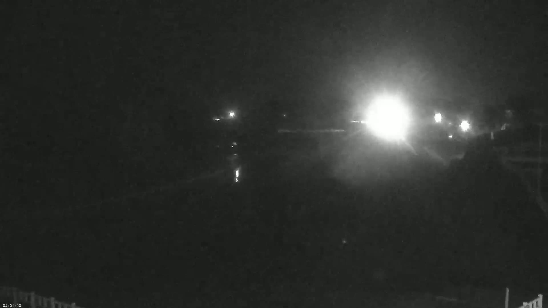 A nighttime, low-light view of a dark body of water with bright headlights from a vehicle approaching in the distance.