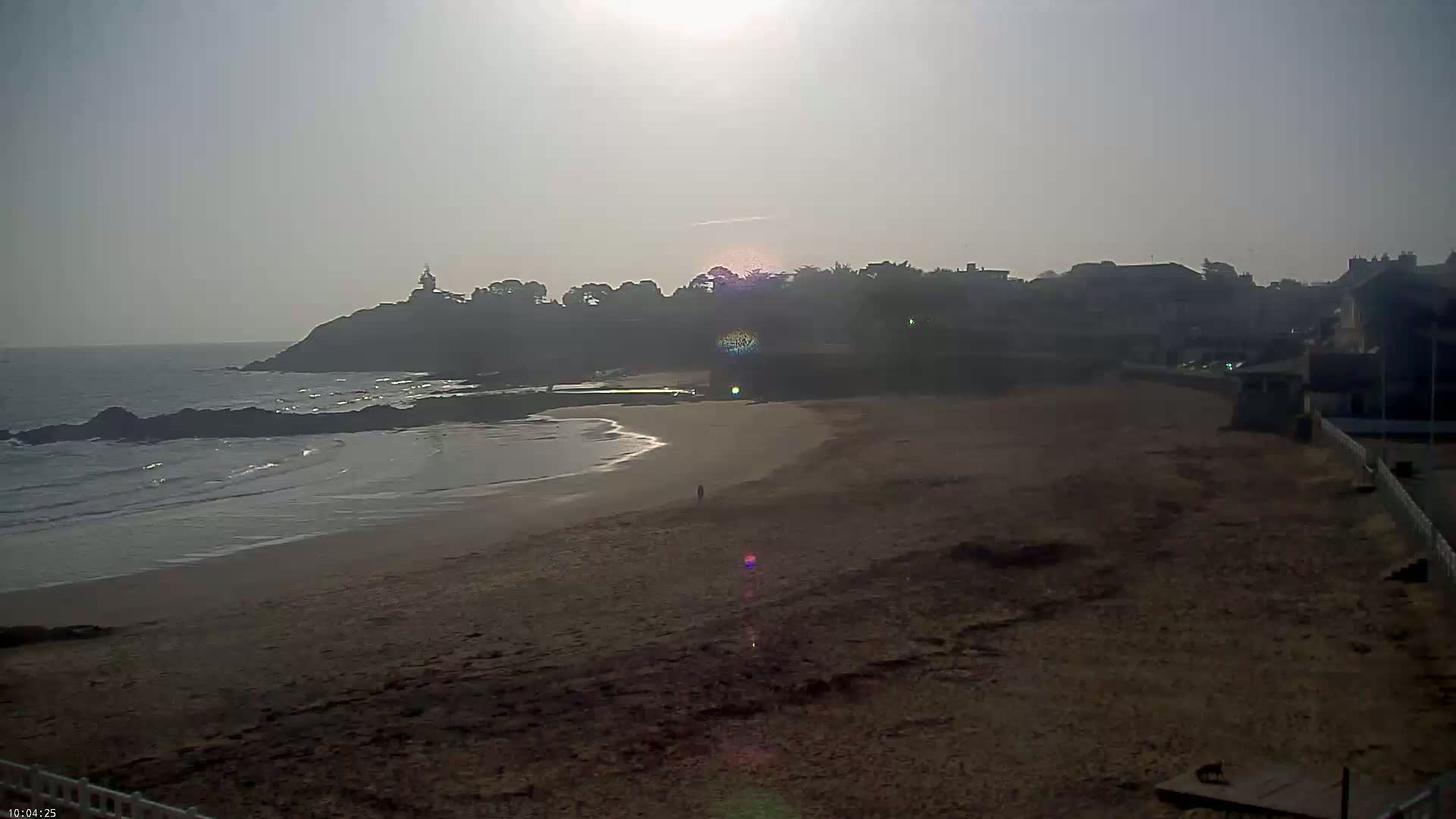 A sandy beach stretches out to a calm ocean under a hazy sun, with a small town and a lighthouse visible on a cliff in the distance.
