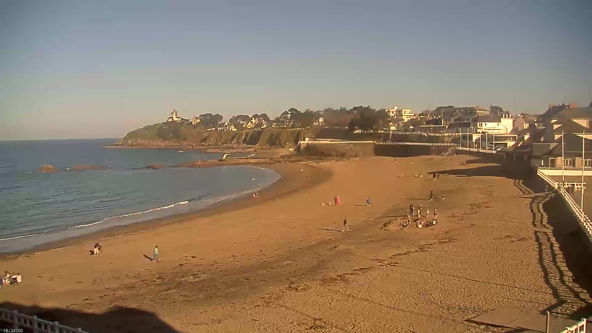 A sandy beach with a few people on it curves gently around a calm ocean, backed by a seaside town under a clear sky.
