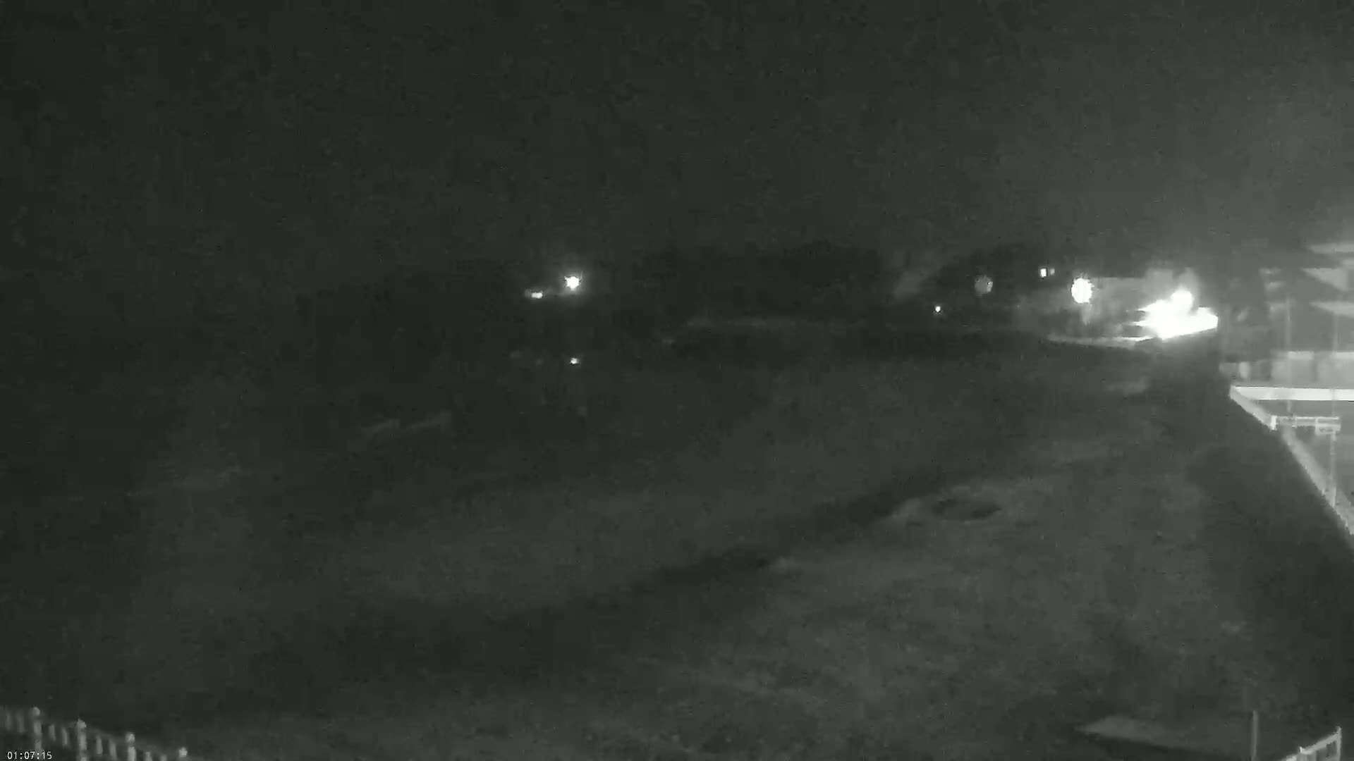 A nighttime, low-light view shows a dark, mostly empty expanse of land with a few distant lights and a portion of a structure visible in the lower right corner.