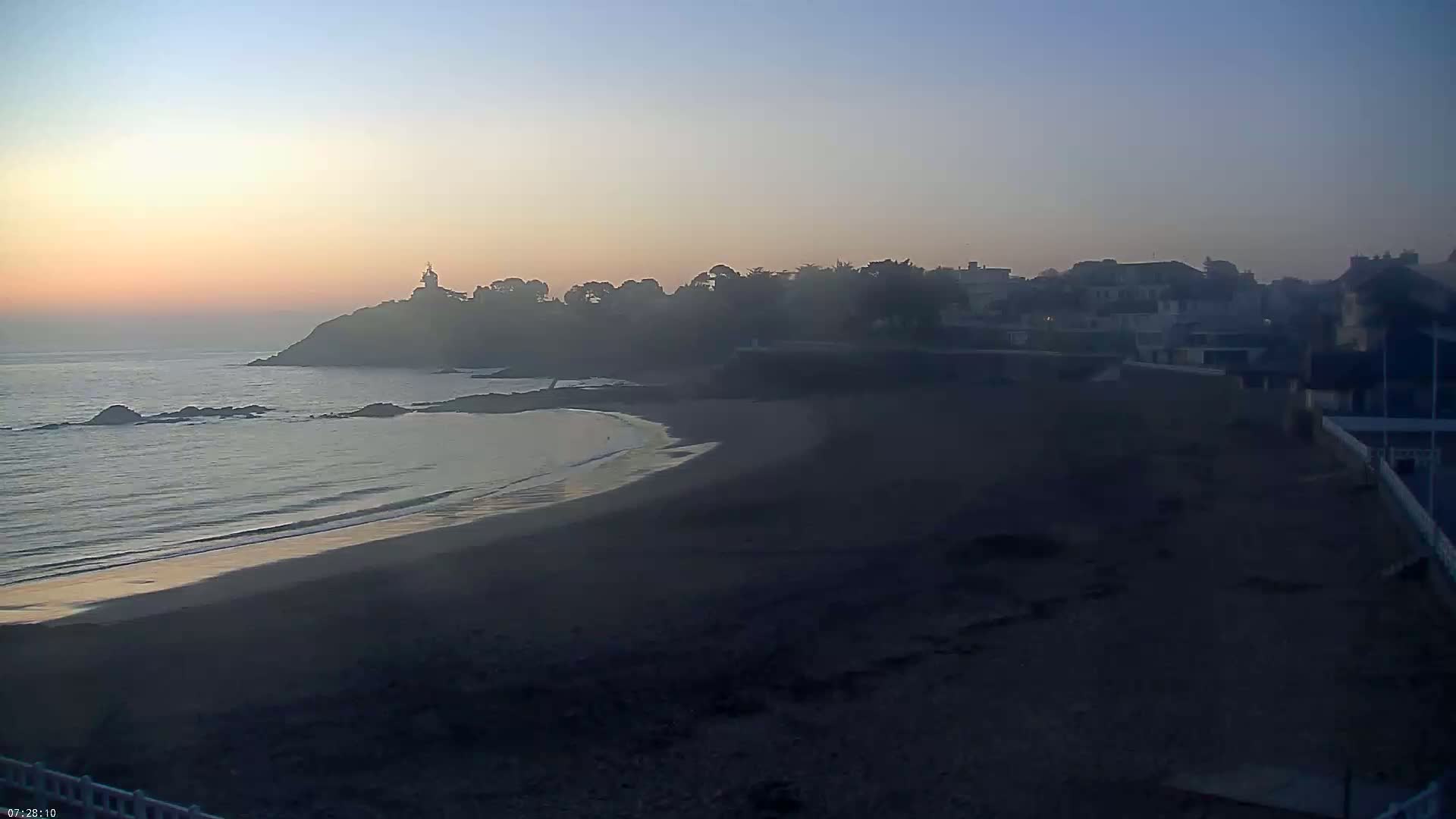 A hazy sunrise illuminates a dark sandy beach and calm ocean waves, with a town and a small lighthouse visible on a distant headland.
