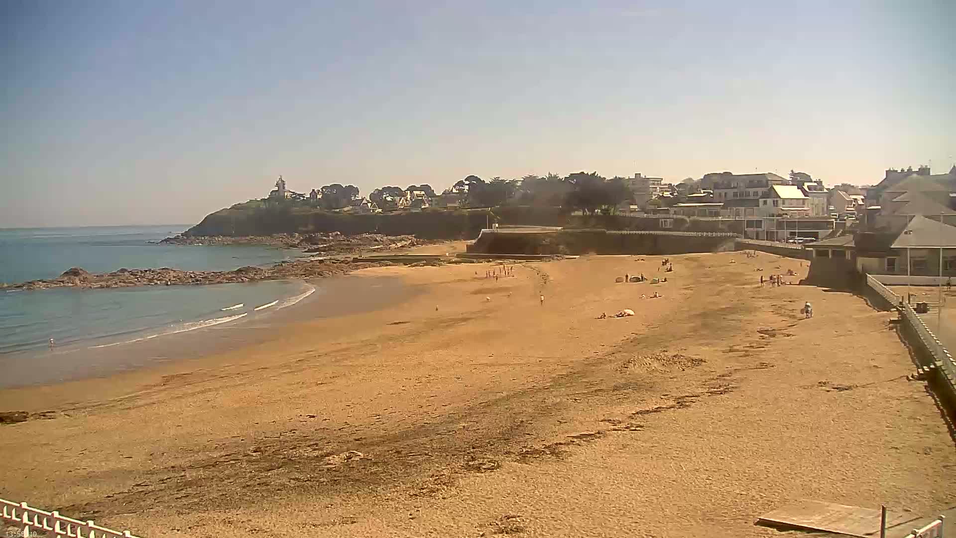 A sandy beach with a few people on it stretches out to a calm ocean under a clear, sunny sky, backed by a coastline of buildings and a small hill with a lighthouse.