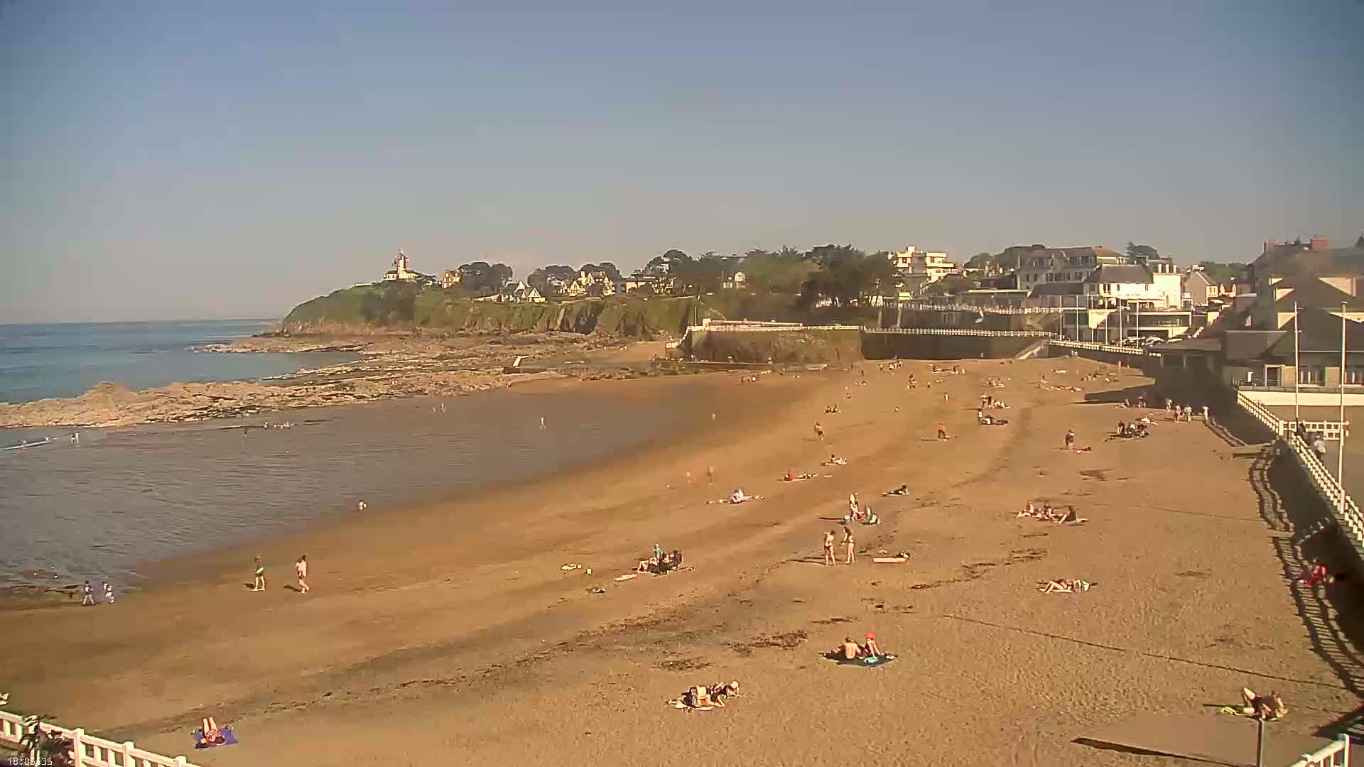 A sandy beach with many people sunbathing and swimming, backed by a town and a rocky headland, under a clear blue sky.