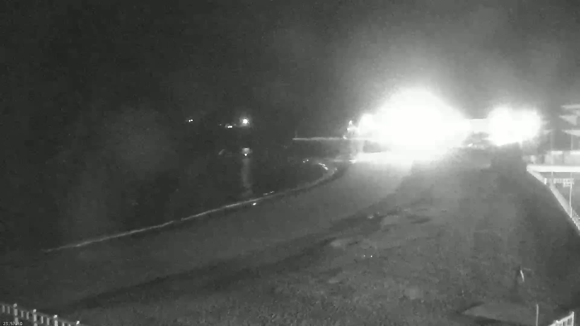 A nighttime, foggy scene shows a curved road with bright headlights approaching from the center, and faint lights in the distance.