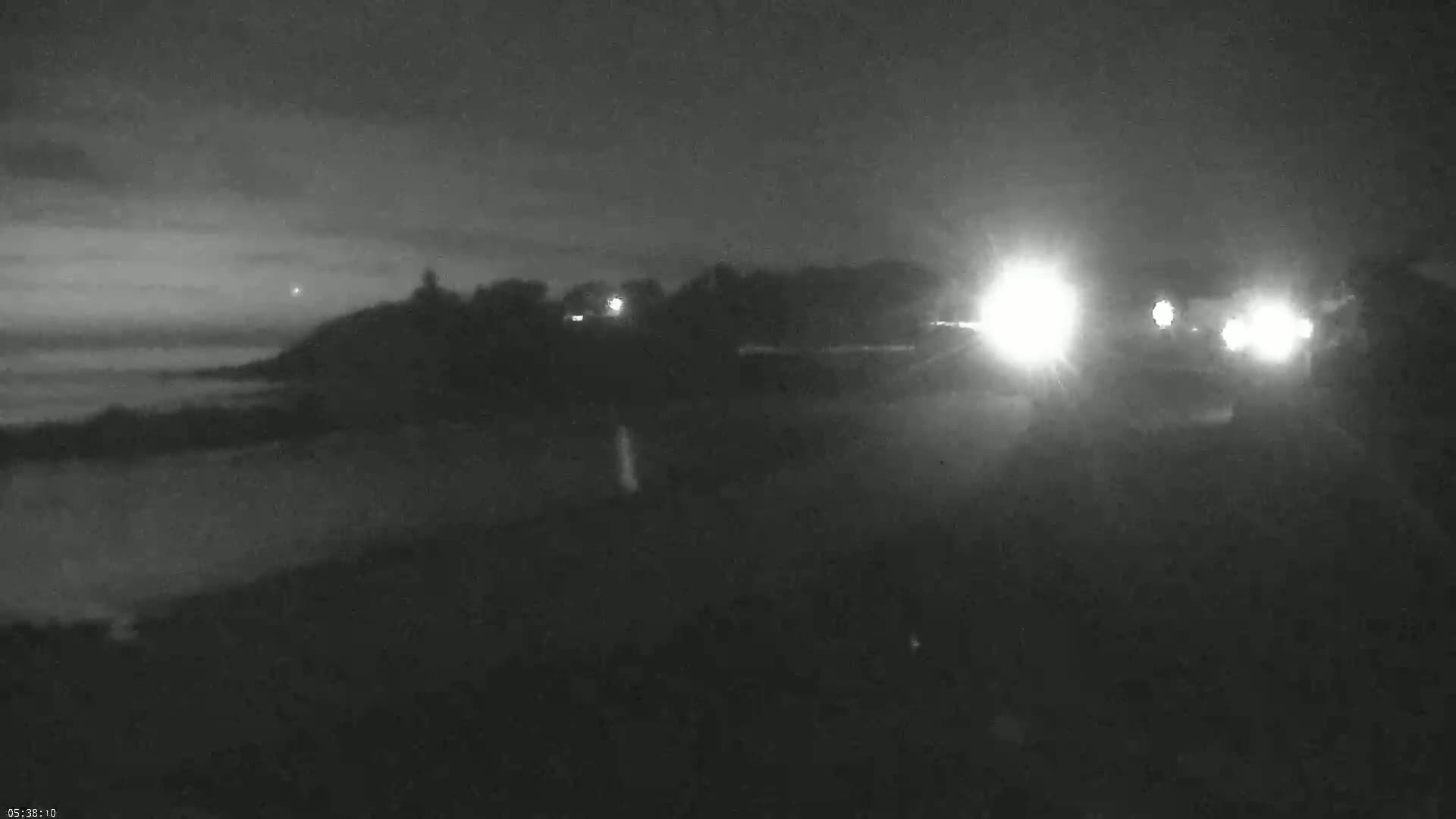 A nighttime, low-light view of a dark shoreline with a body of water, land, and distant vehicle headlights.