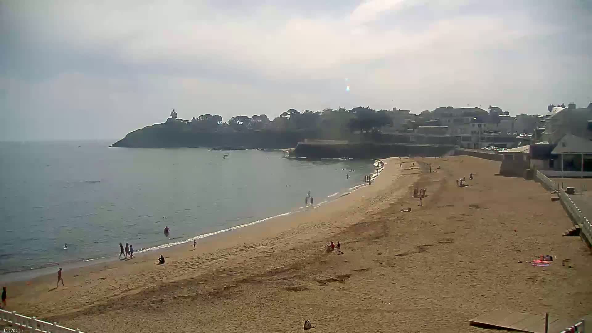 A sandy beach with scattered people on it, stretches alongside calm ocean water towards a small town nestled against a rocky headland under a mostly cloudy sky.