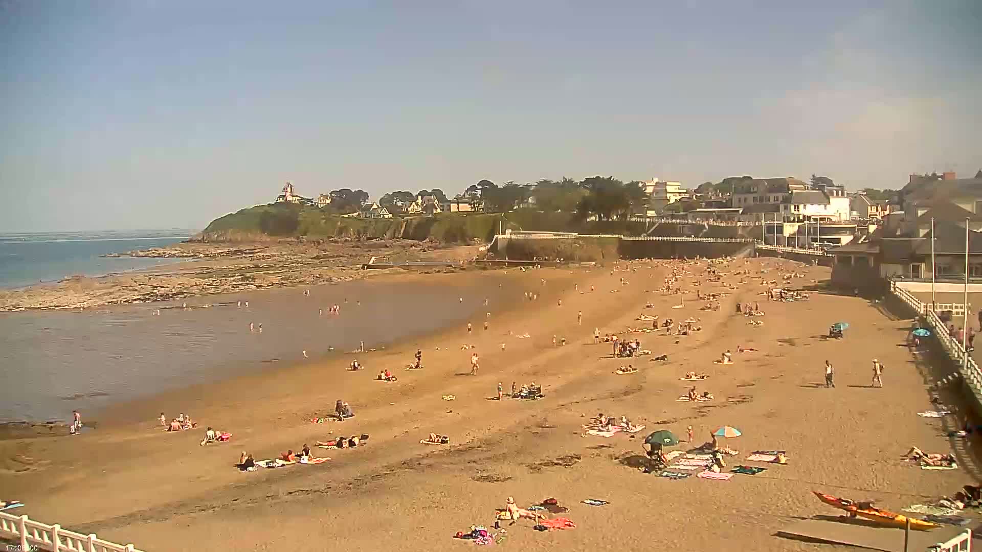 A sandy beach with many people sunbathing and swimming, situated next to a calm body of water and a seaside town under a clear, sunny sky.