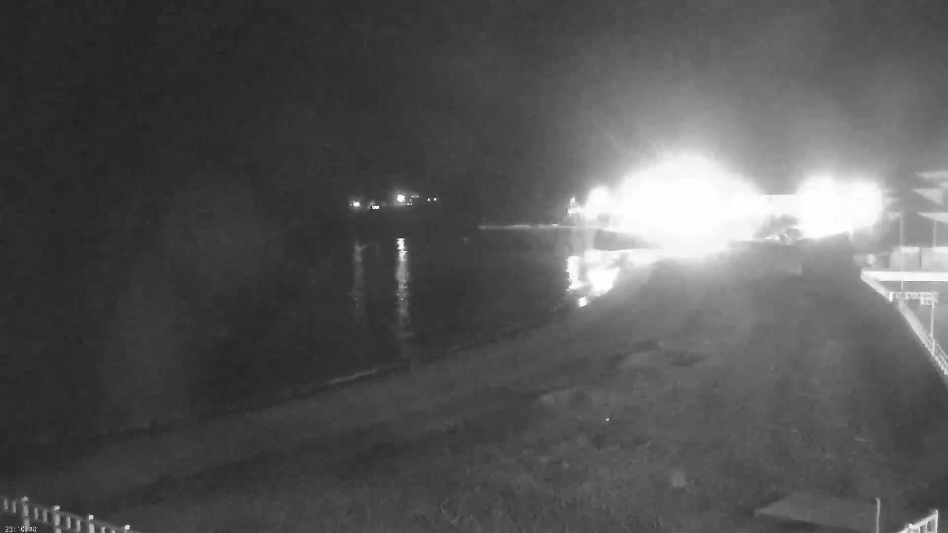 A nighttime, low-light view shows a bright light source near a body of water, with a dark shoreline and a building partially visible in the lower right corner.