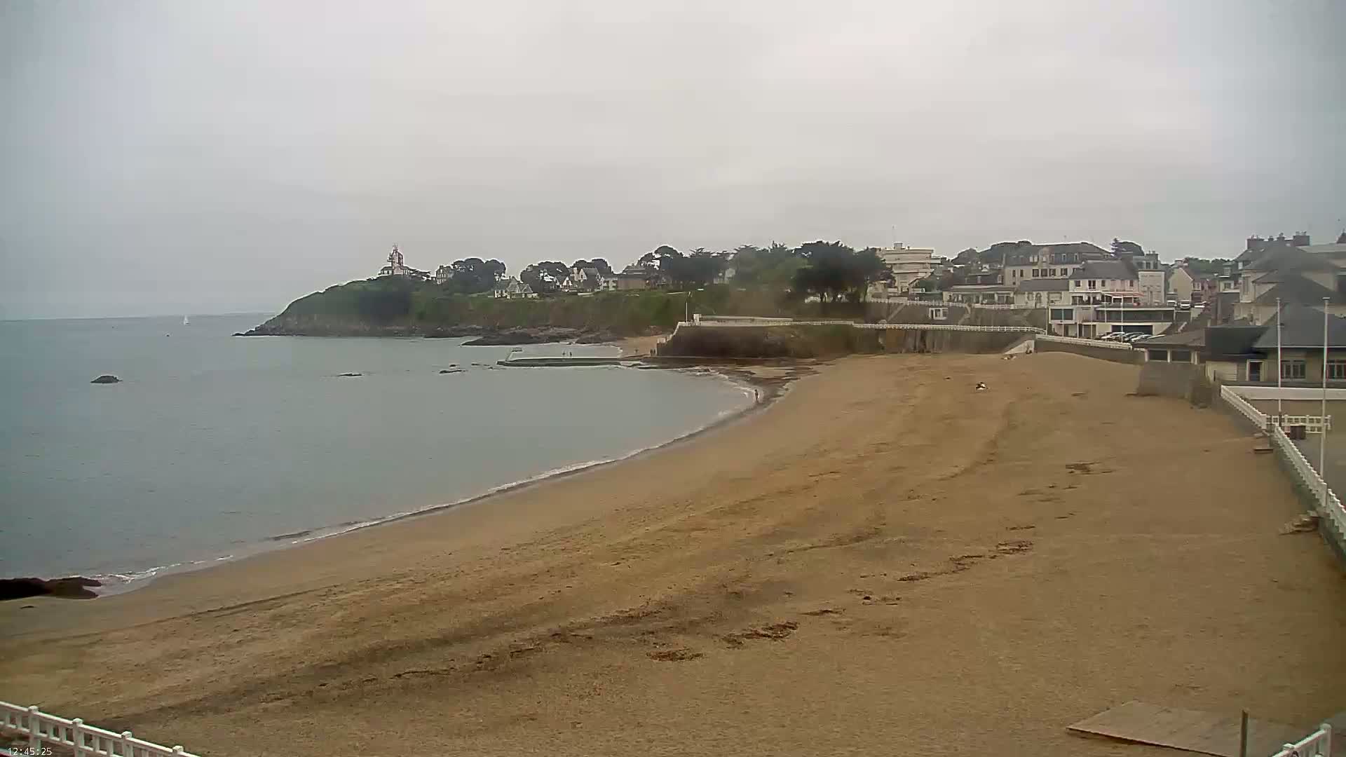 A sandy beach curves around a calm, grey sea toward a small town on a cloudy day.