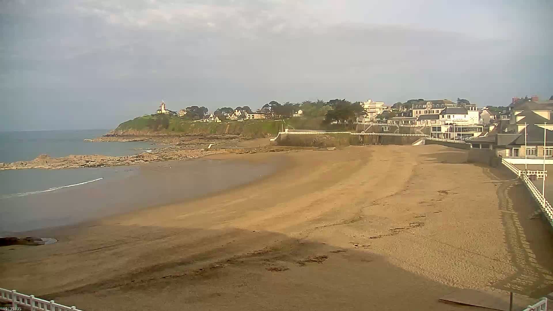 A sandy beach curves toward a calm ocean, with a collection of buildings and a lighthouse visible on a cliff in the distance under an overcast sky.