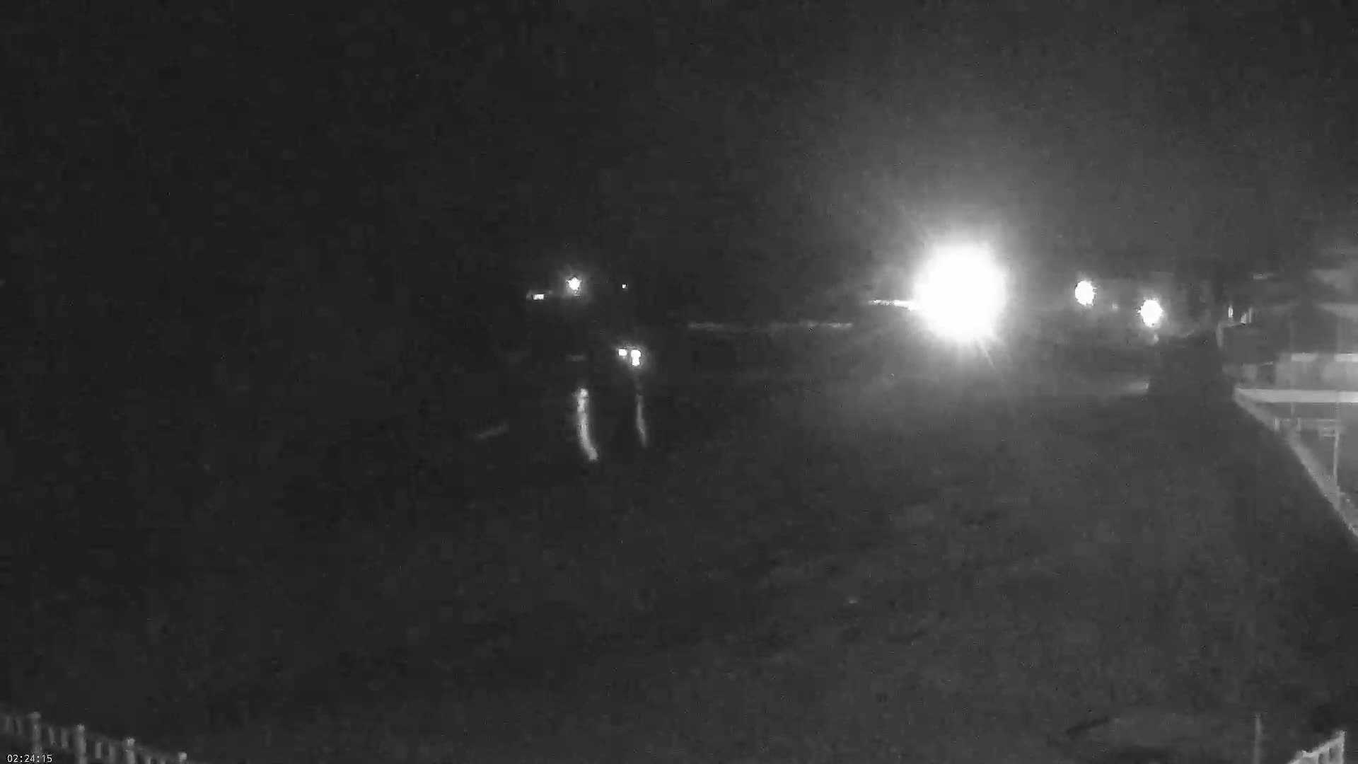 A nighttime, low-light image shows a dark, possibly sandy or paved area with vehicle headlights in the distance and a structure to the right.