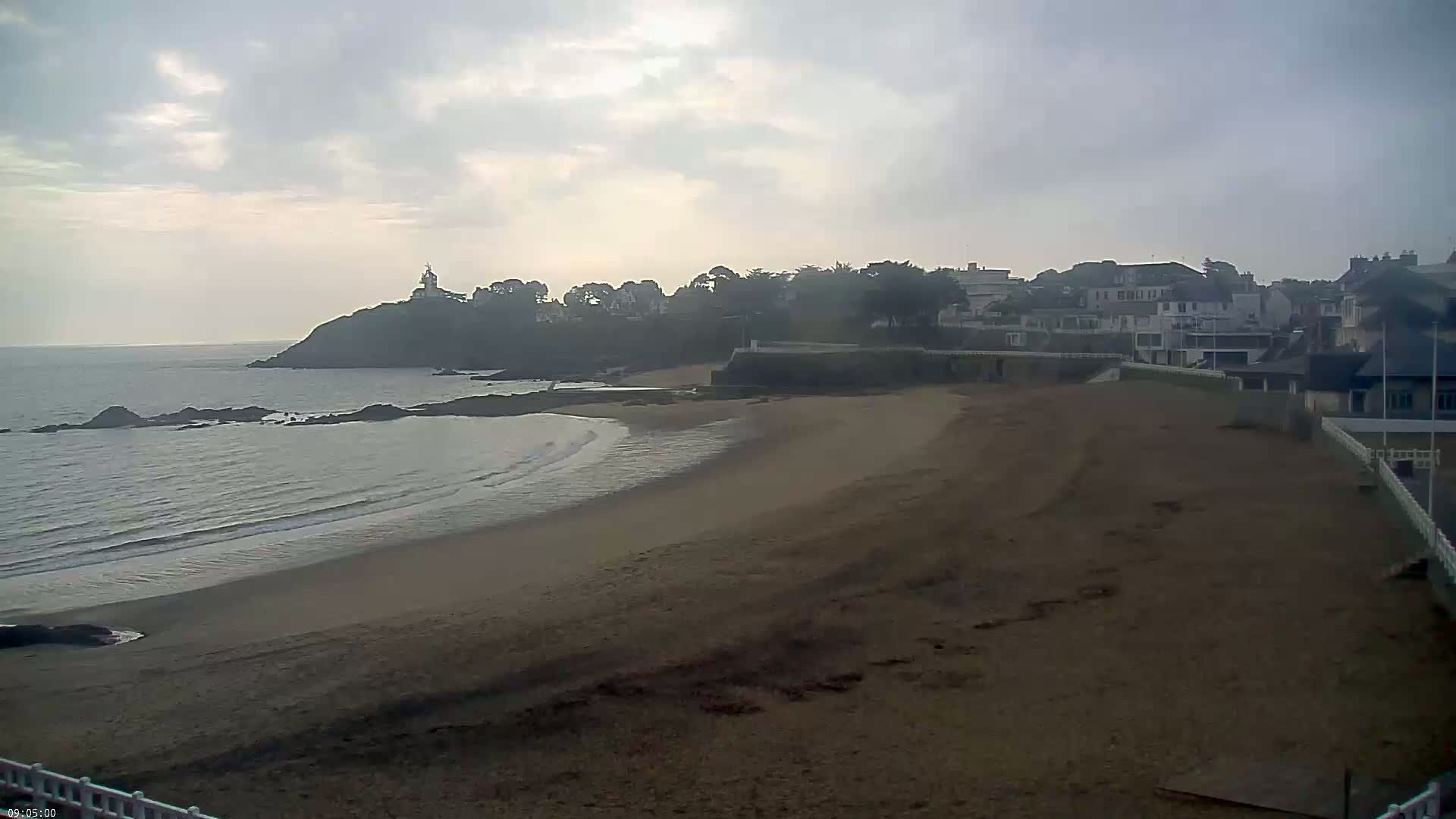 A sandy beach curves around a calm ocean, bordered by a town with buildings and a small lighthouse on a nearby headland, all under an overcast sky.
