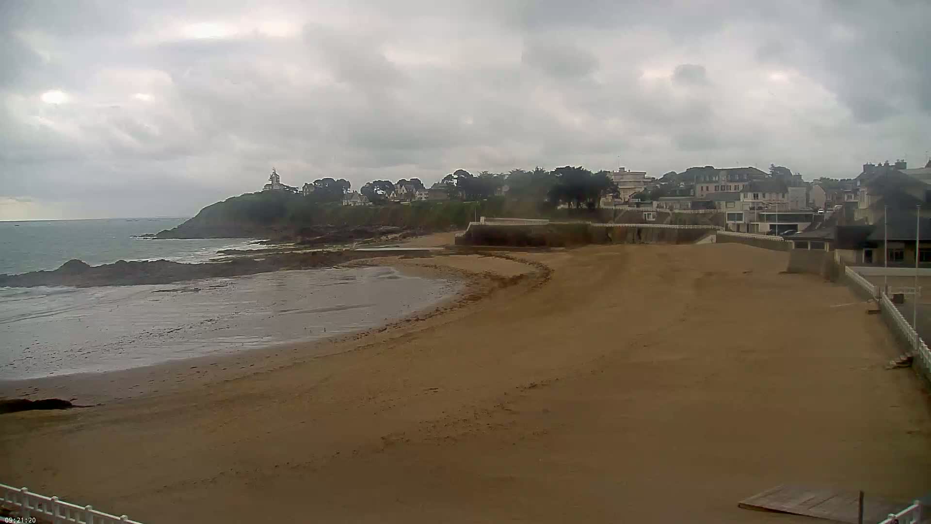 A sandy beach curves towards a calm ocean under an overcast sky, with a small town and a lighthouse visible on a nearby headland.