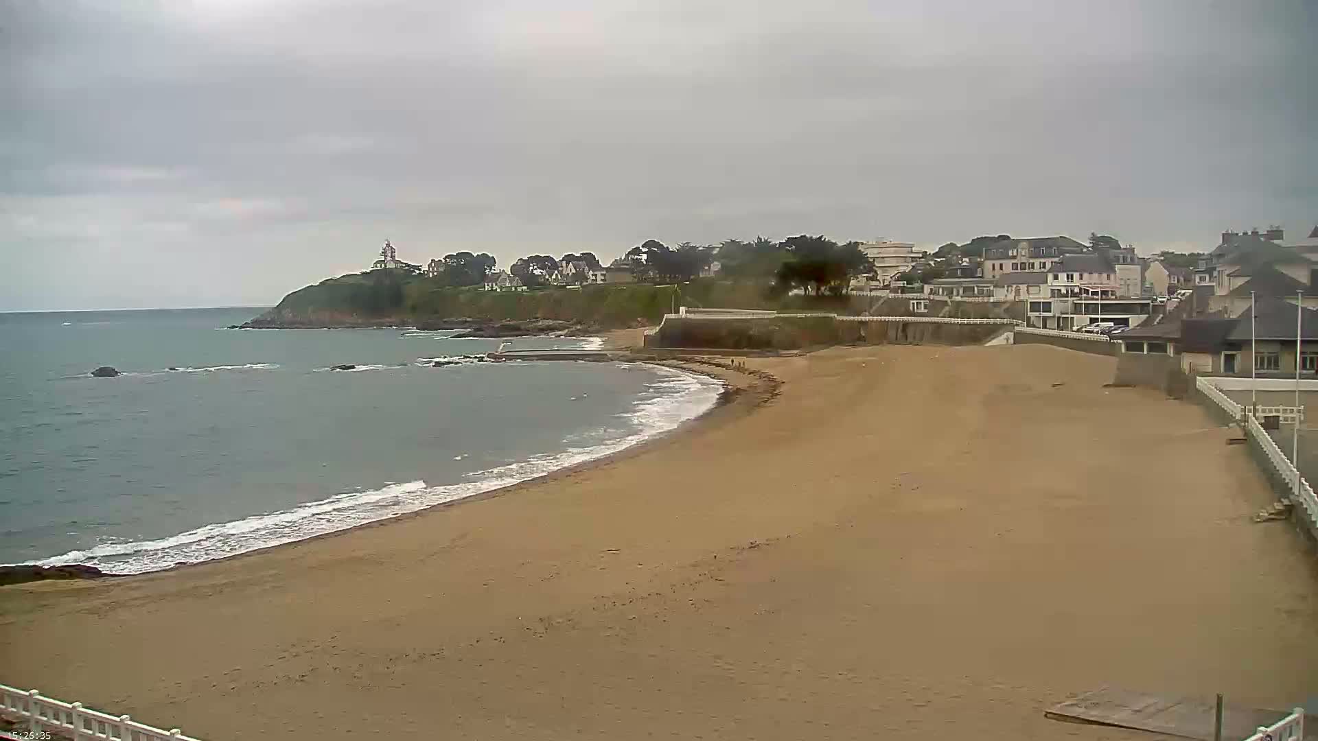 A sandy beach curves gently around a calm, grey sea, backed by a seaside town under an overcast sky.