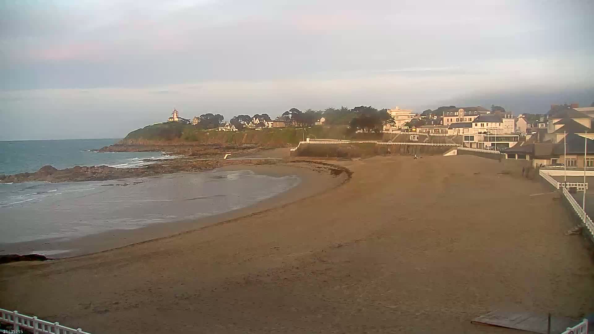 A sandy beach curves gently around a rocky point, with calm ocean water, a few buildings and a lighthouse visible in the distance under an overcast sky.