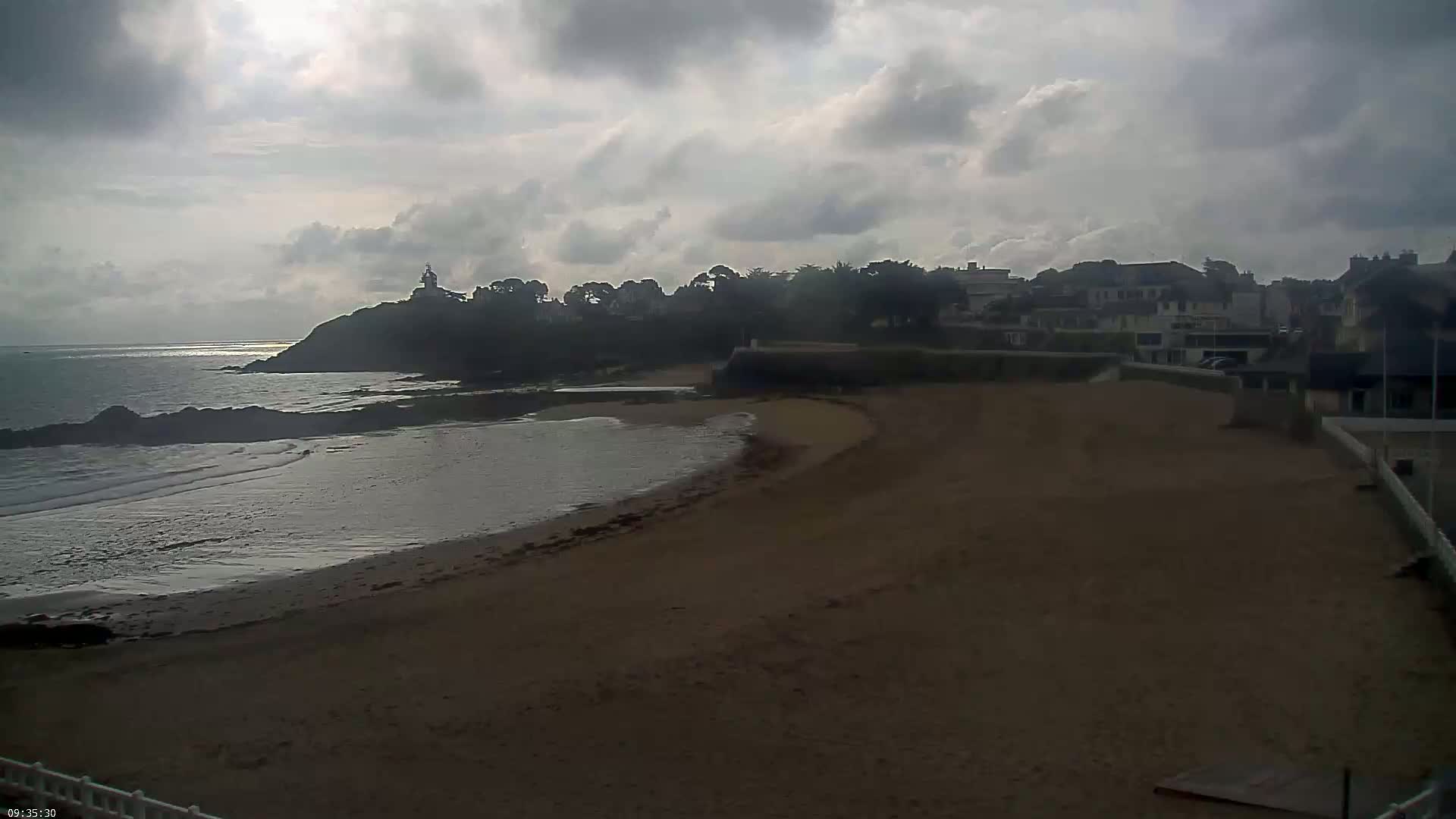 A sandy beach curves around a calm bay, with a town and a lighthouse silhouetted against a cloudy sky.