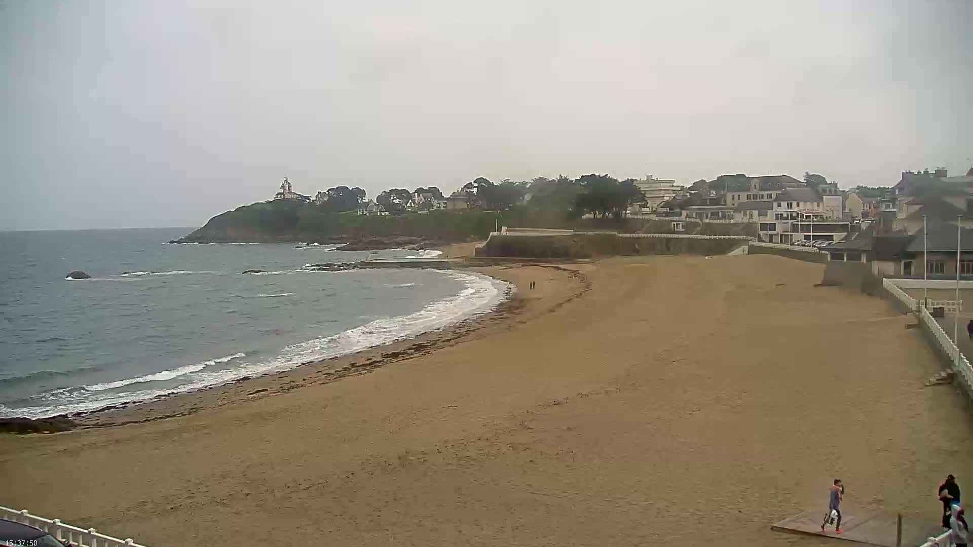 A sandy beach curves around a small cove, with a few people visible on the sand and buildings and a lighthouse visible on the cliffs in the background, under an overcast sky.
