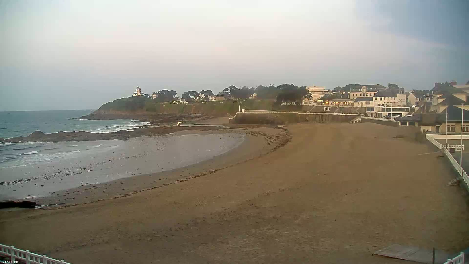A sandy beach stretches towards a town on a cloudy day, with a calm ocean and a small lighthouse visible in the distance.
