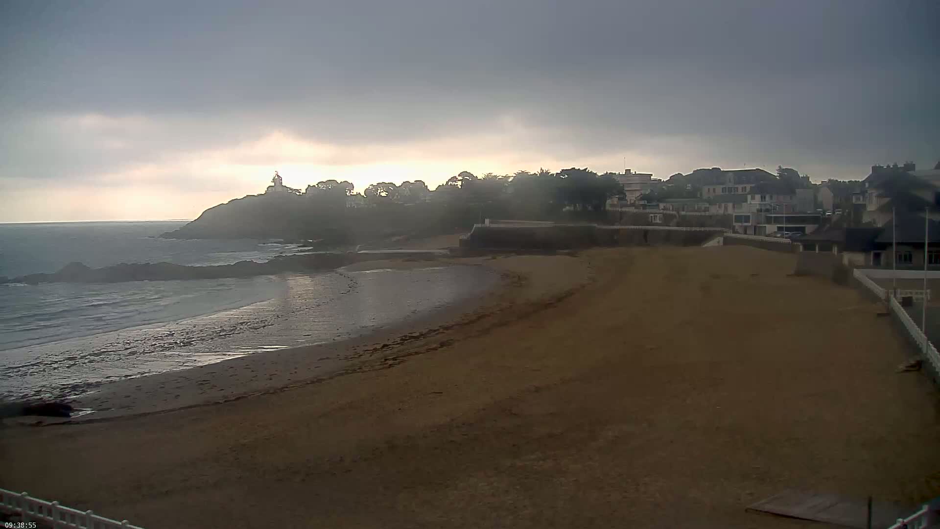 A sandy beach curves around a calm ocean, with buildings and a lighthouse visible on a hill in the distance under an overcast sky.