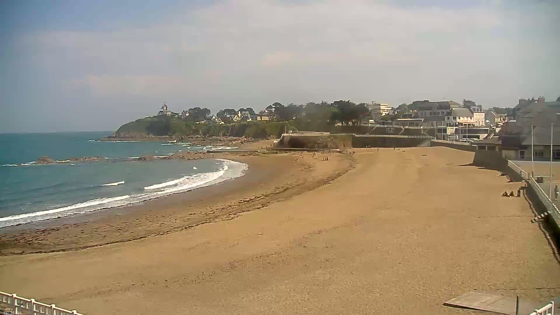 A sandy beach stretches along a calm ocean under a partly cloudy sky, with a small town and a lighthouse visible on a cliff in the background.
