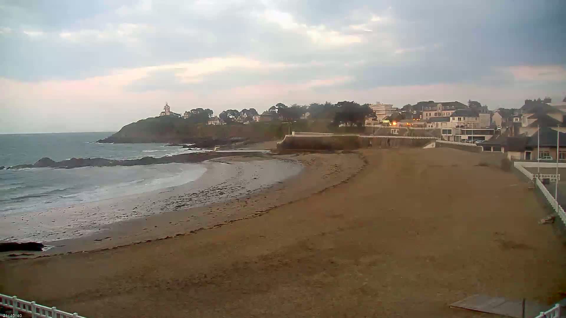 A sandy beach curves around a rocky shoreline, meeting a calm ocean under an overcast sky, with a village visible on a hill in the background.