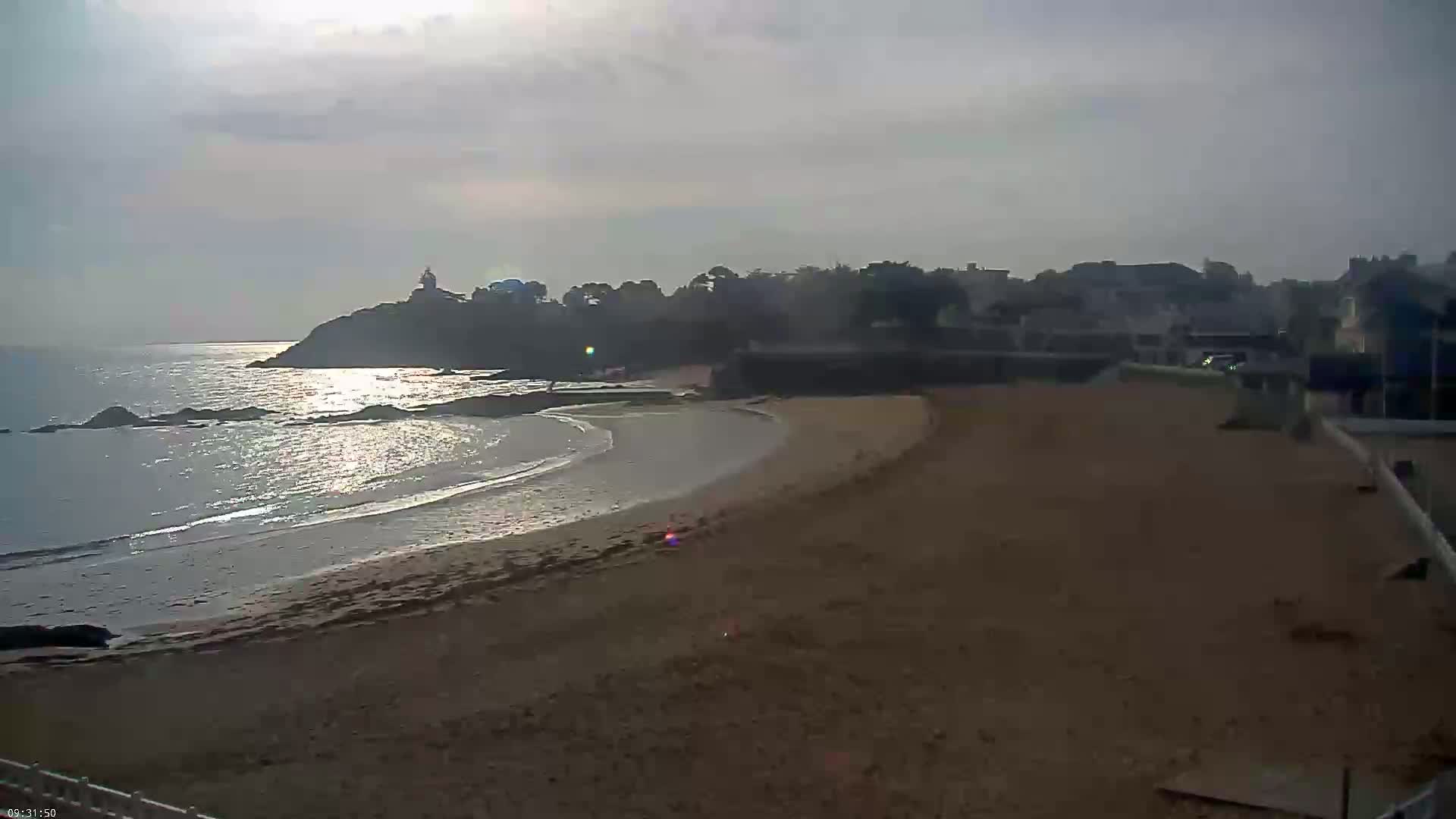 A sandy beach curves around a rocky point, with calm waves lapping the shore under a mostly cloudy sky.