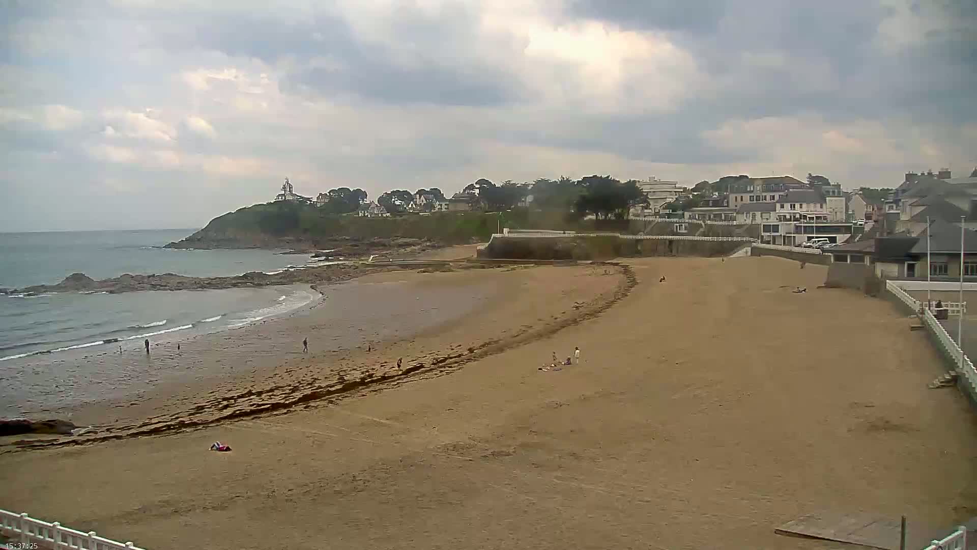 A sandy beach with a few people on it stretches out to a calm ocean under a cloudy sky, backed by a town on a small cliff.