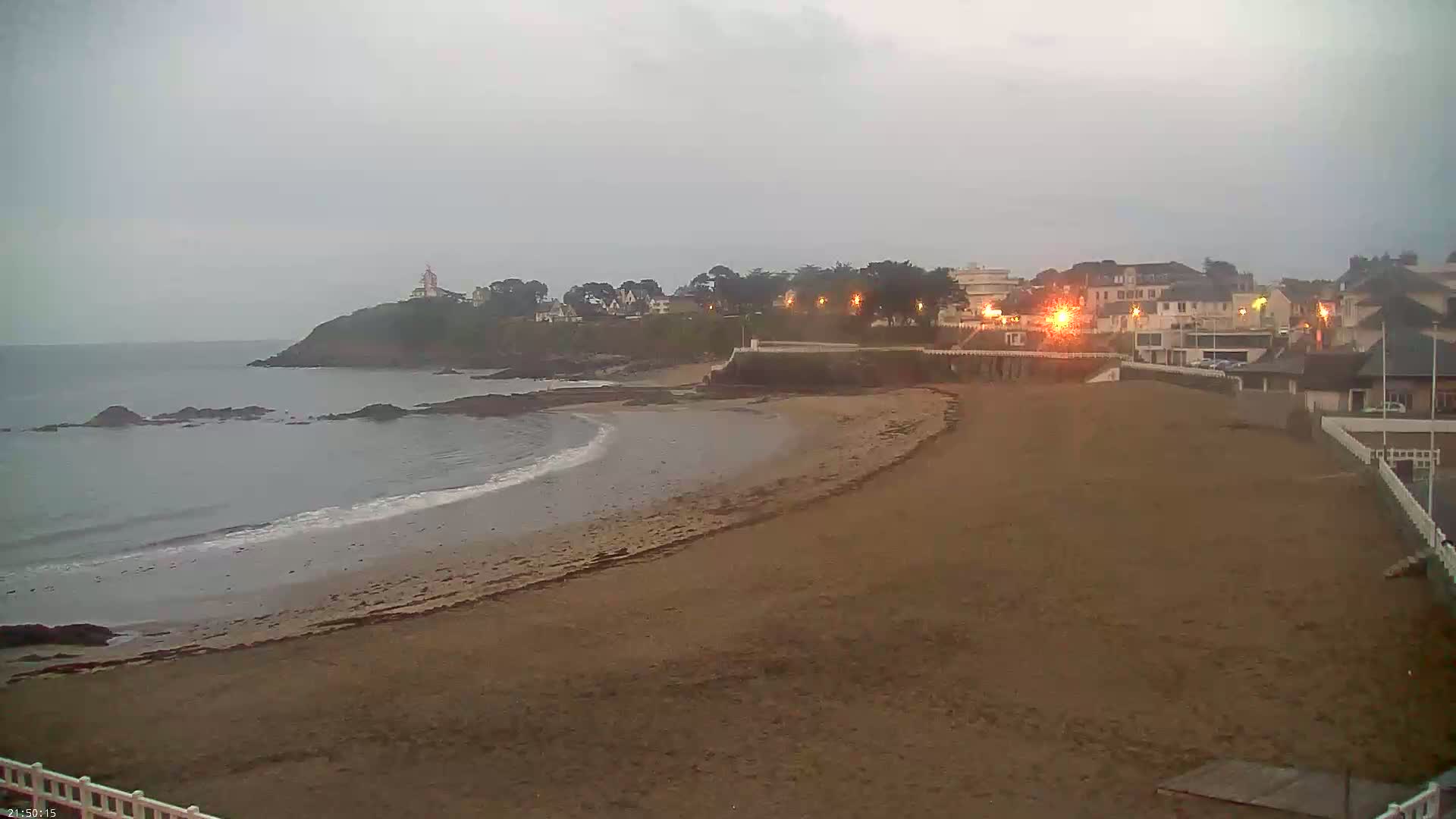 A sandy beach curves gently around a rocky point, with calm waves lapping the shore under an overcast sky, and a town with lights visible in the distance.
