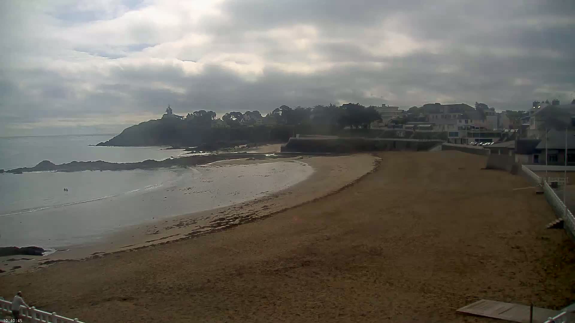 A sandy beach curves around a calm bay, backed by a line of buildings and a rocky headland under a cloudy sky.