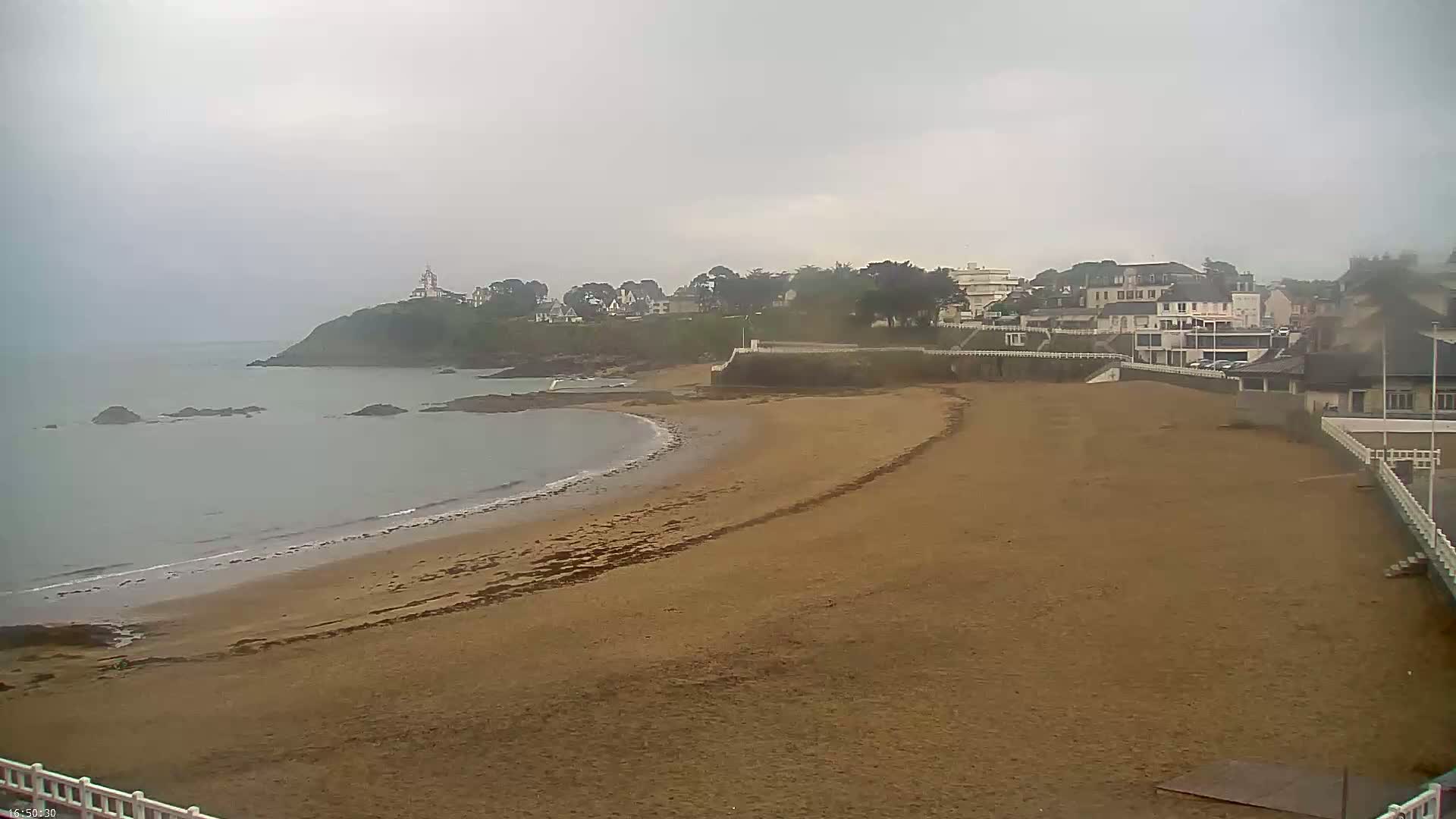 A sandy beach curves around a calm, gray ocean under an overcast sky, with a small town and a lighthouse visible on a cliff in the background.