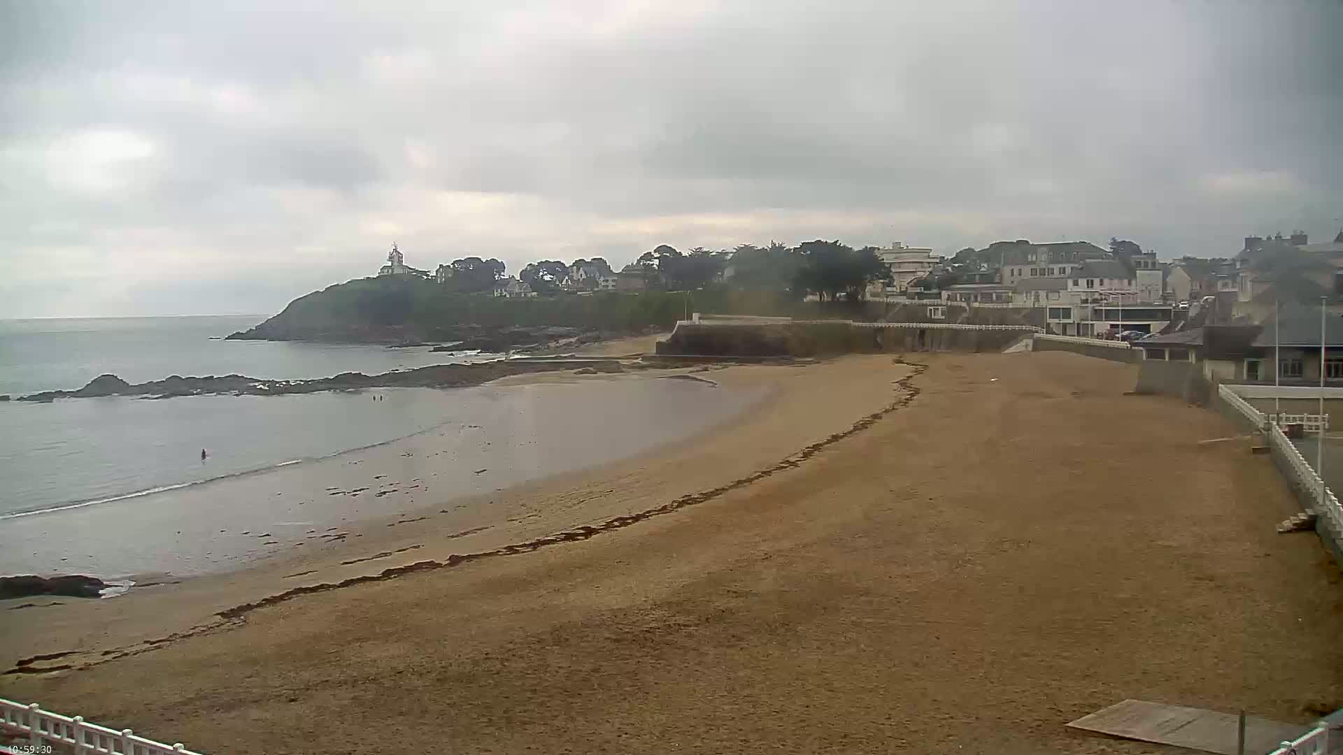 A sandy beach curves along a calm sea under an overcast sky, with a small town and a lighthouse visible on a cliff in the distance.