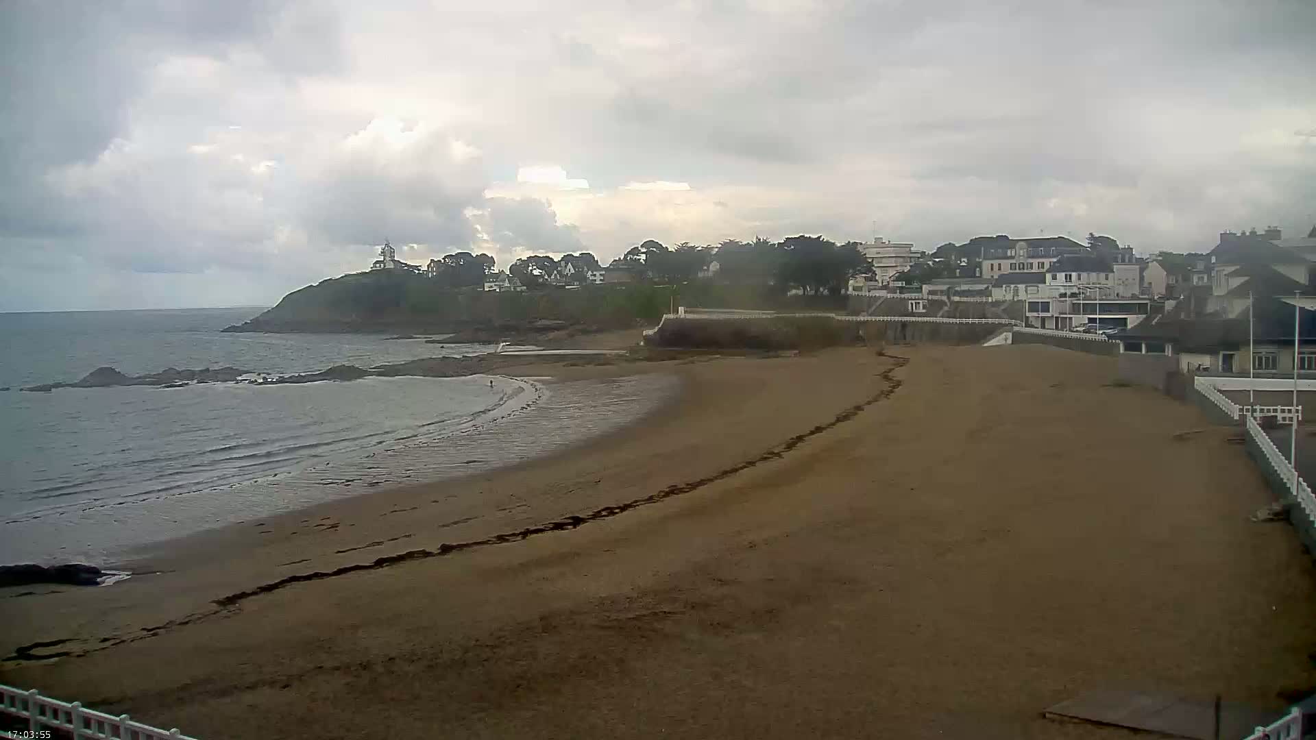 A sandy beach curves around a calm bay under a cloudy sky, with buildings and a small lighthouse visible on a distant headland.