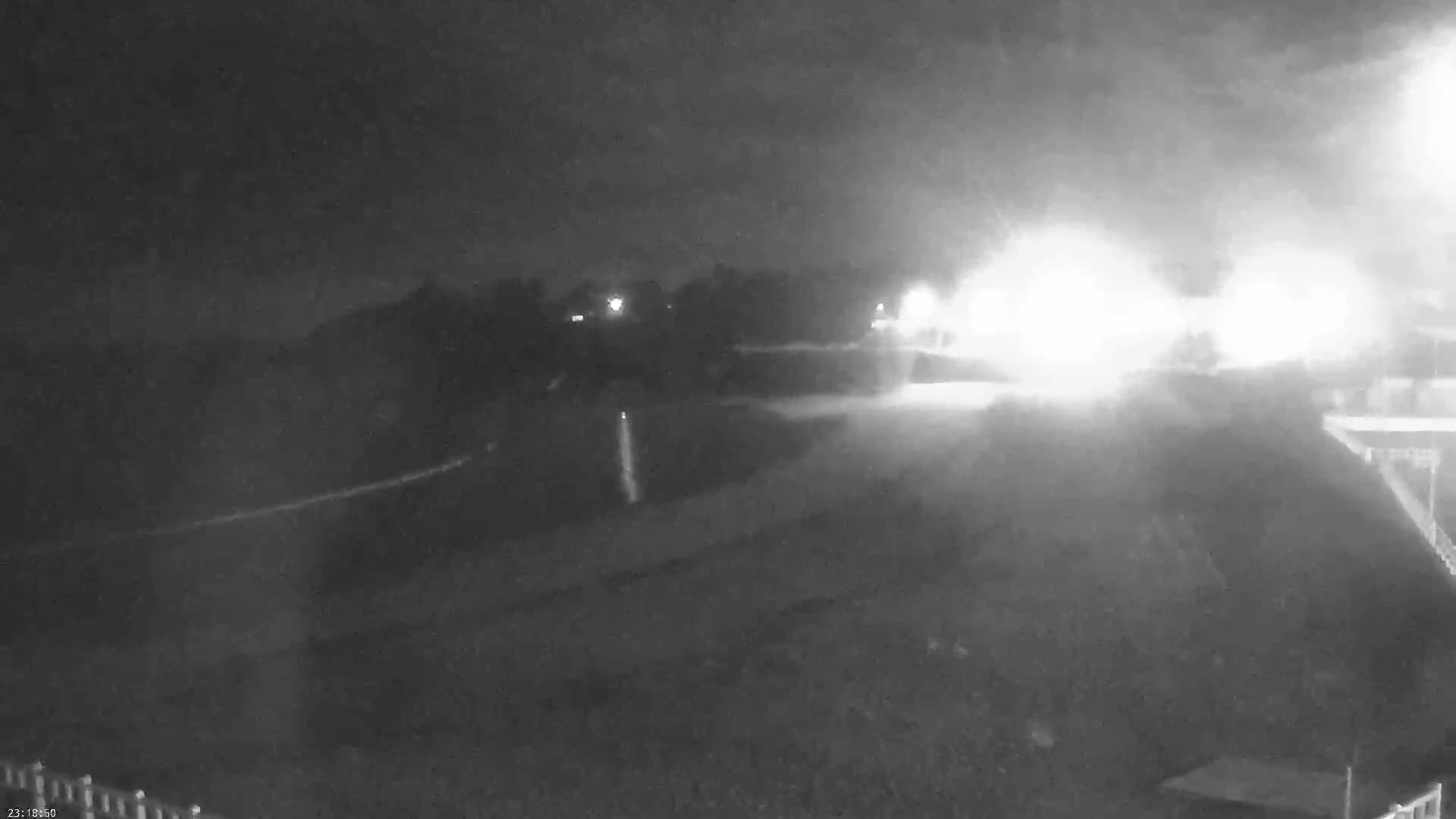 A nighttime, overcast view shows a dark road with bright lights in the distance, surrounded by dark trees and buildings.