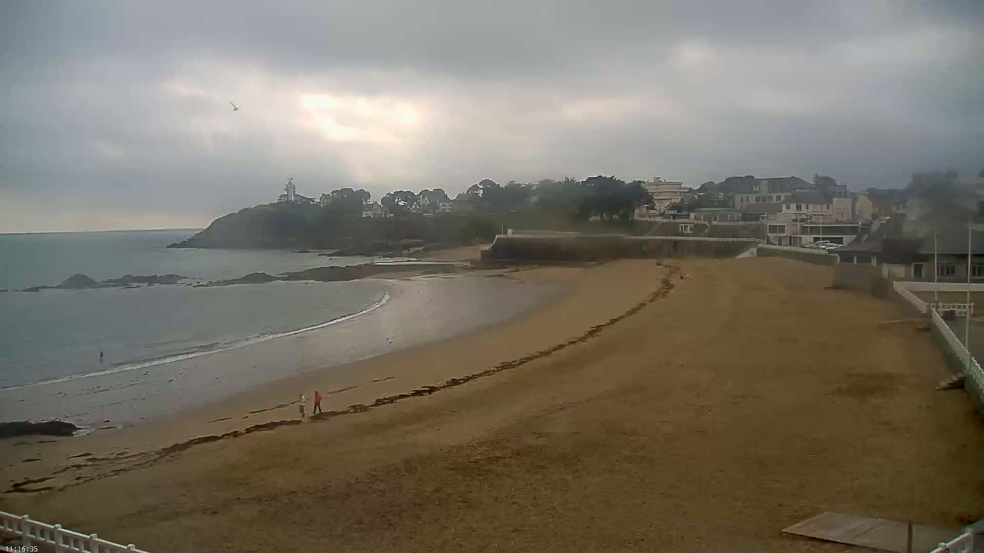 A sandy beach curves along a coastline under an overcast sky, with a few people walking near the water's edge and buildings visible on the headland.