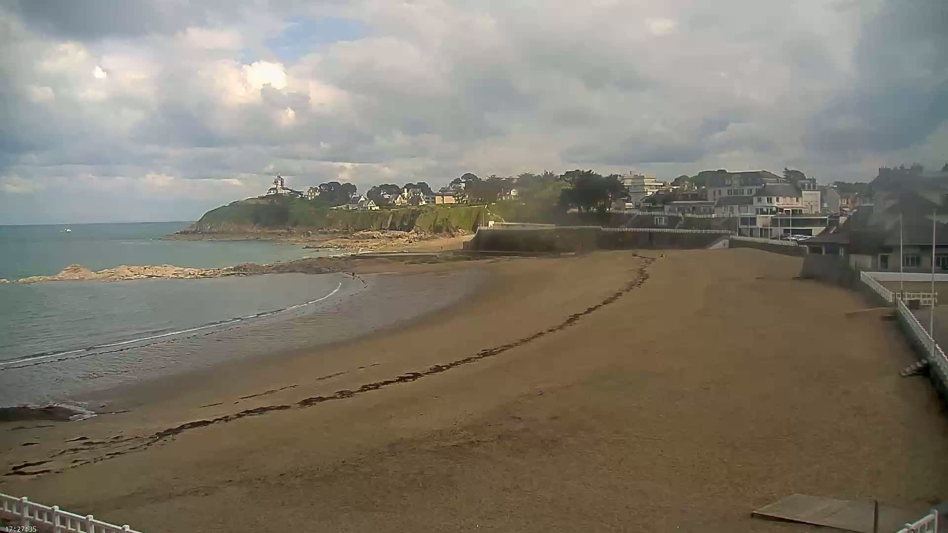 A sandy beach curves gently into a calm sea under a partly cloudy sky, with a small town and a lighthouse visible on a nearby cliff.