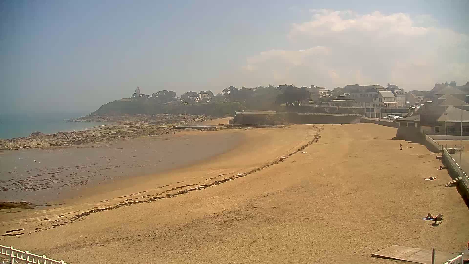 A sandy beach stretches from a rocky shoreline toward a line of buildings and a hill under a mostly sunny sky.