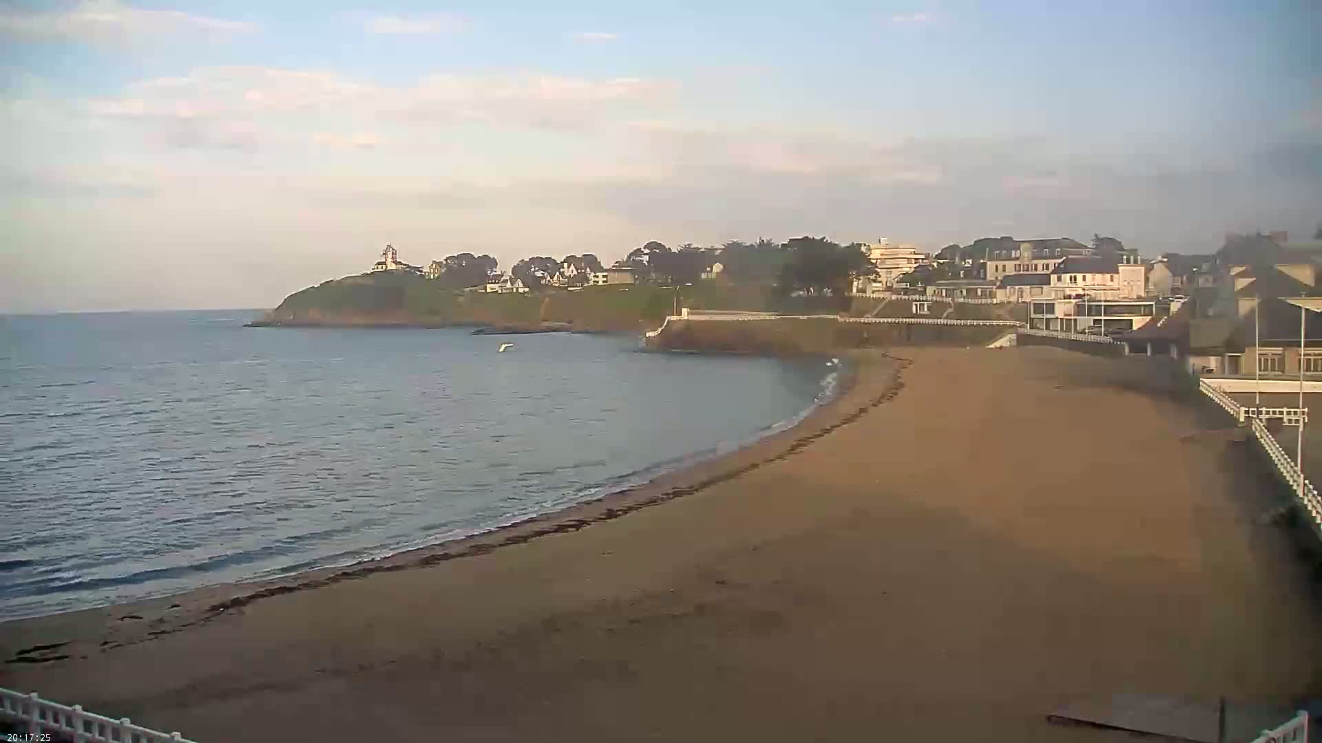 A sandy beach curves gently to meet calm ocean waves lapping the shore, with buildings and a lighthouse visible on a nearby cliff under a mostly clear sky.