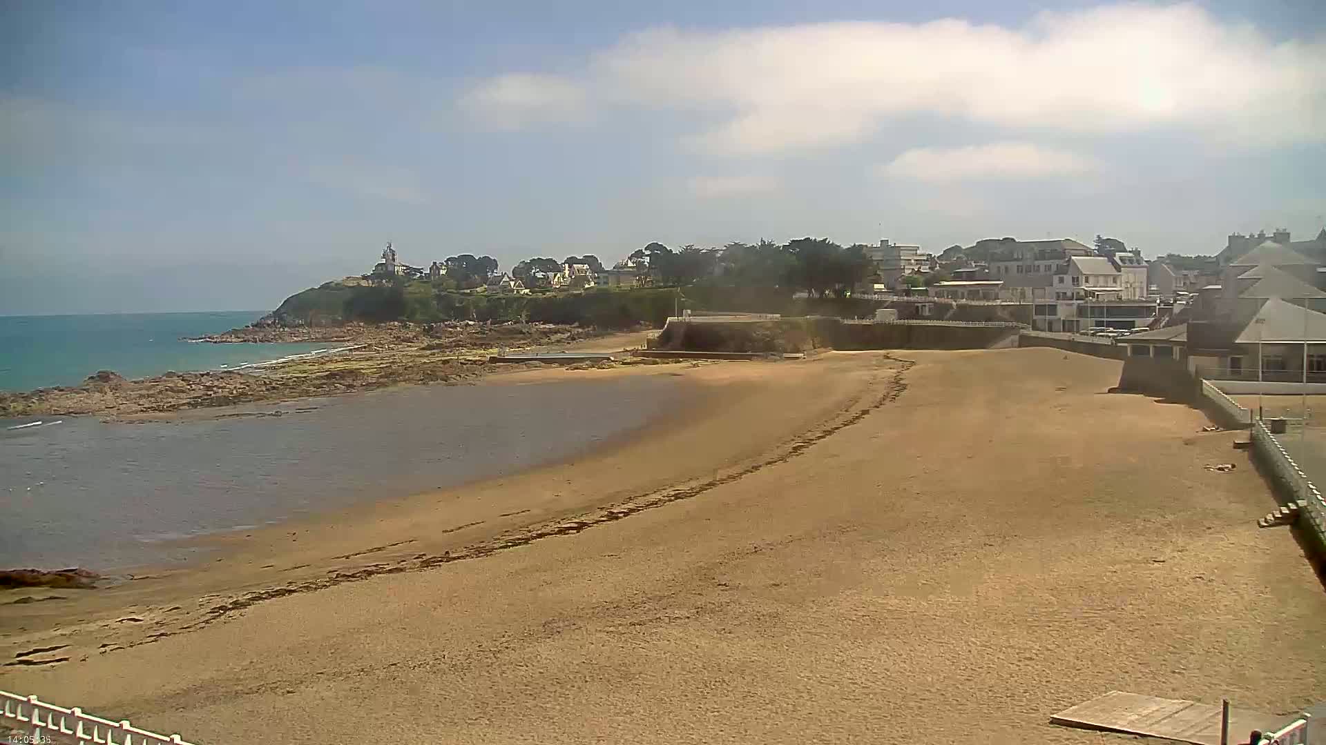A sandy beach stretches out to a calm body of water under a mostly sunny sky, with buildings and a rocky coast visible in the background.