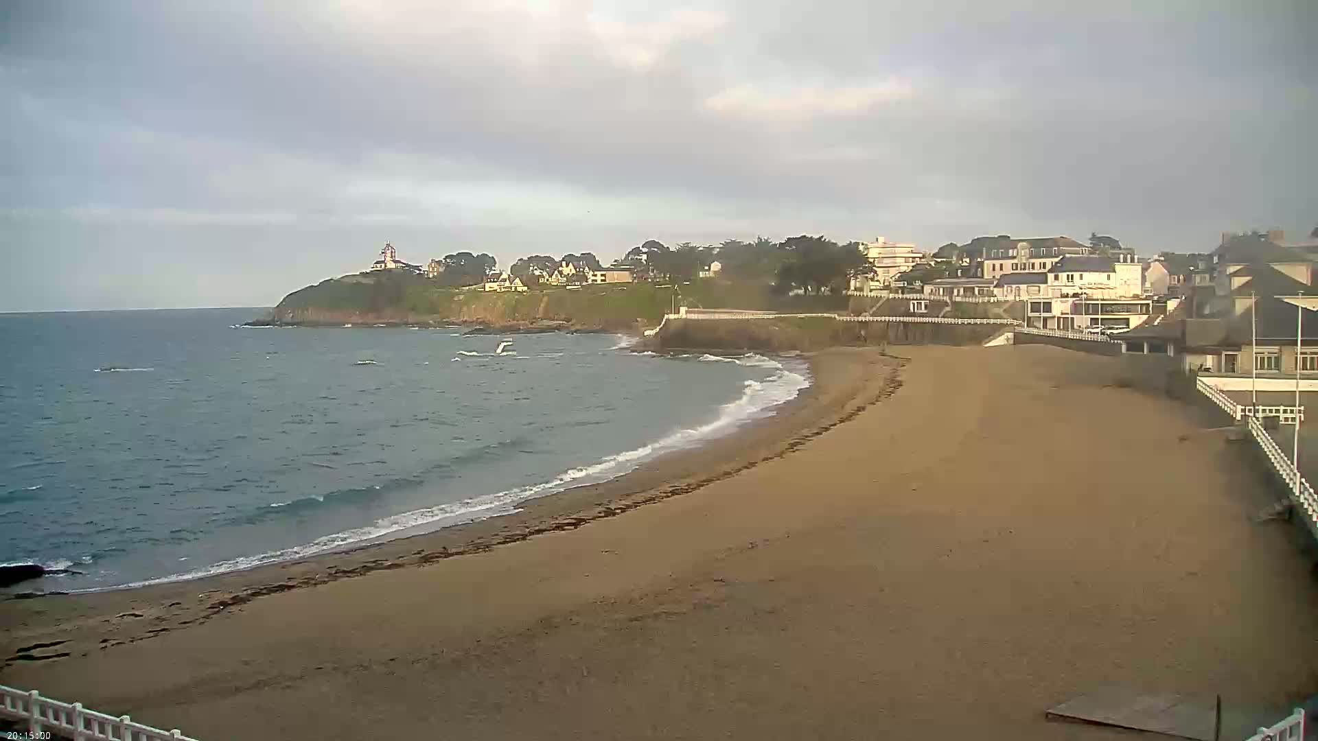 A sandy beach curves along a coastline under a cloudy sky, with buildings and a lighthouse visible on a cliff in the distance, and gentle waves lapping the shore.