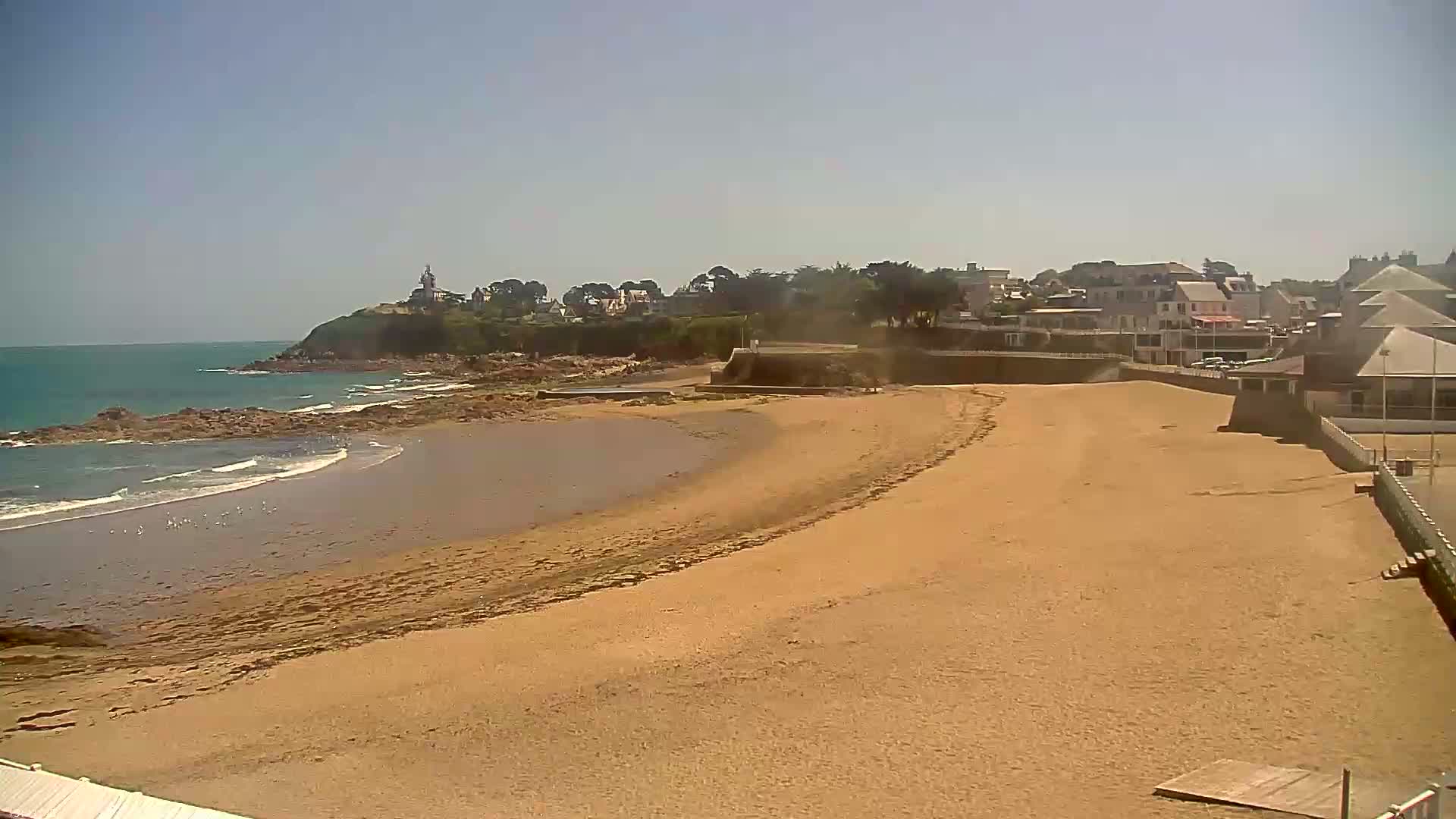 A sandy beach stretches towards a small town under a clear sky, with a calm ocean visible beyond.