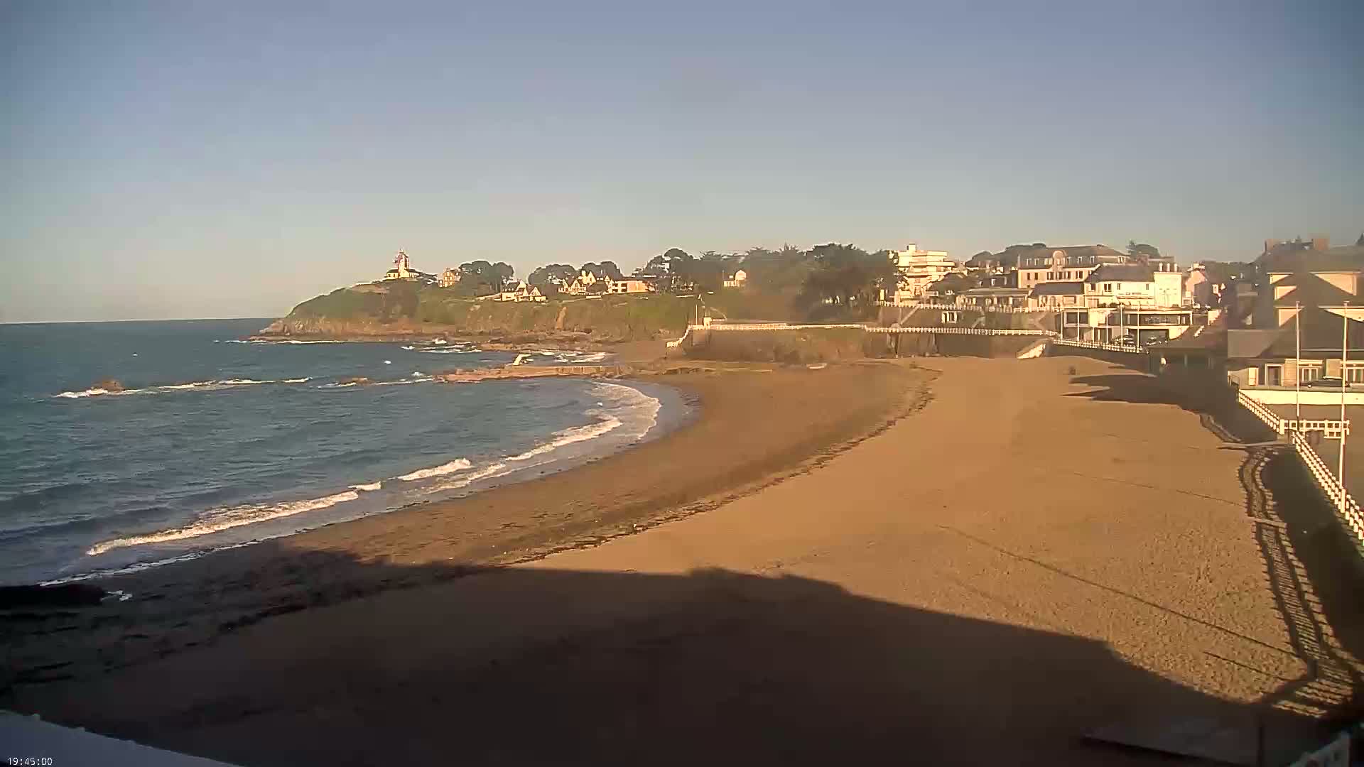 A sandy beach curves gently toward a calm ocean, with houses and a lighthouse perched on a cliff in the background under a clear sky.