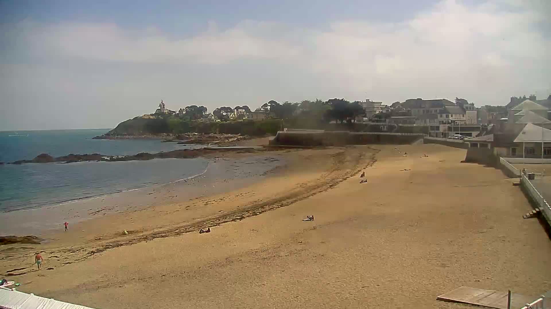 A sandy beach stretches out to a calm ocean under a partly cloudy sky, with a small town and a rocky headland visible in the background.