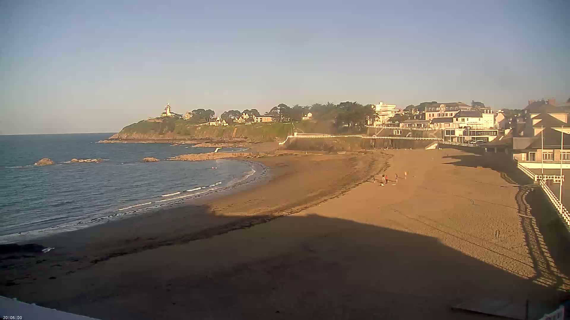 A sandy beach curves around a rocky point, with calm ocean water, a few people, and buildings perched on a cliff in the background under a clear, bright sky.