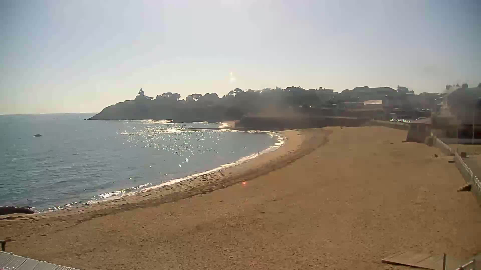A sandy beach curves gently around a calm bay, with a small town and a lighthouse visible on a distant headland under a clear, sunny sky.