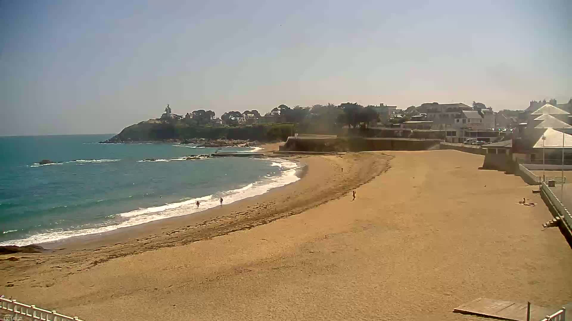 A sandy beach curves gently around a calm, blue ocean under a clear, sunny sky, with buildings and a small lighthouse visible in the distance.