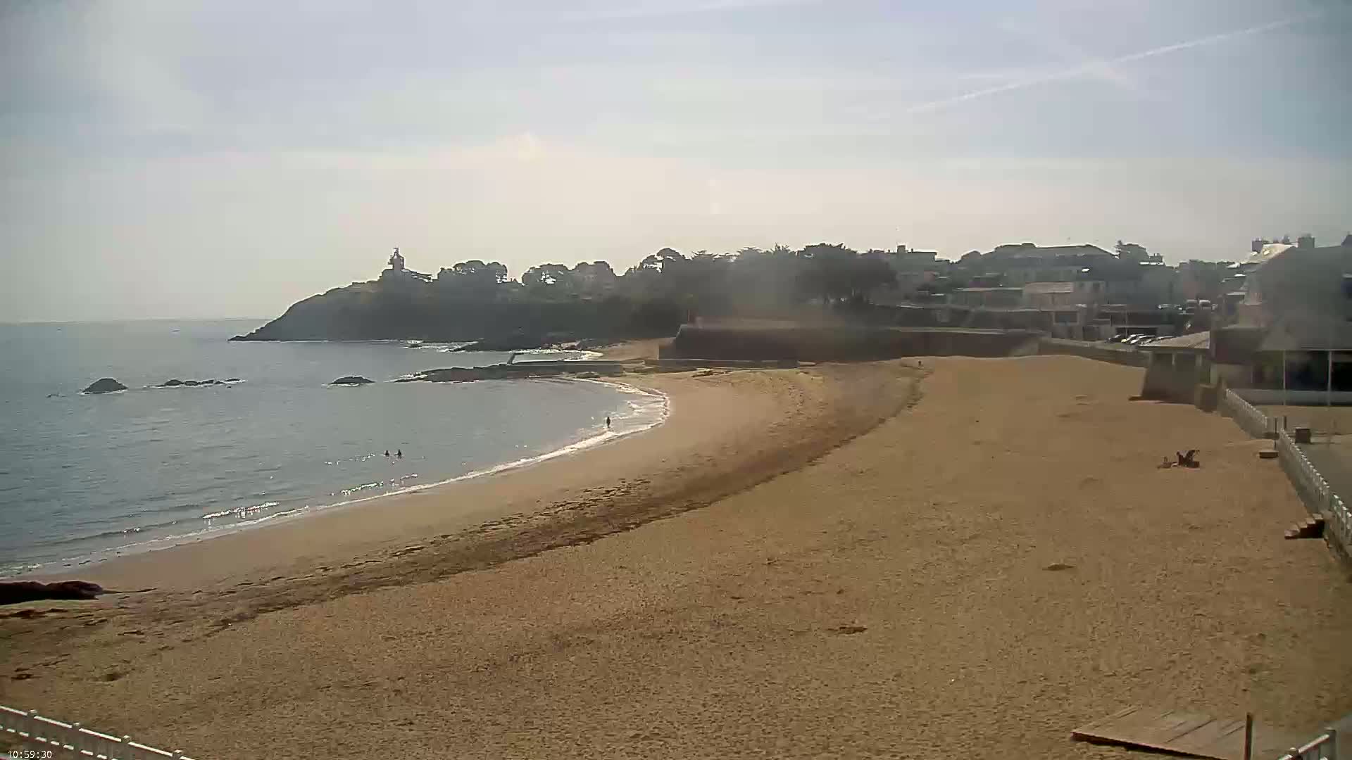A sandy beach curves around a calm ocean, with a few people visible in the water and on the shore, under a mostly sunny sky.
