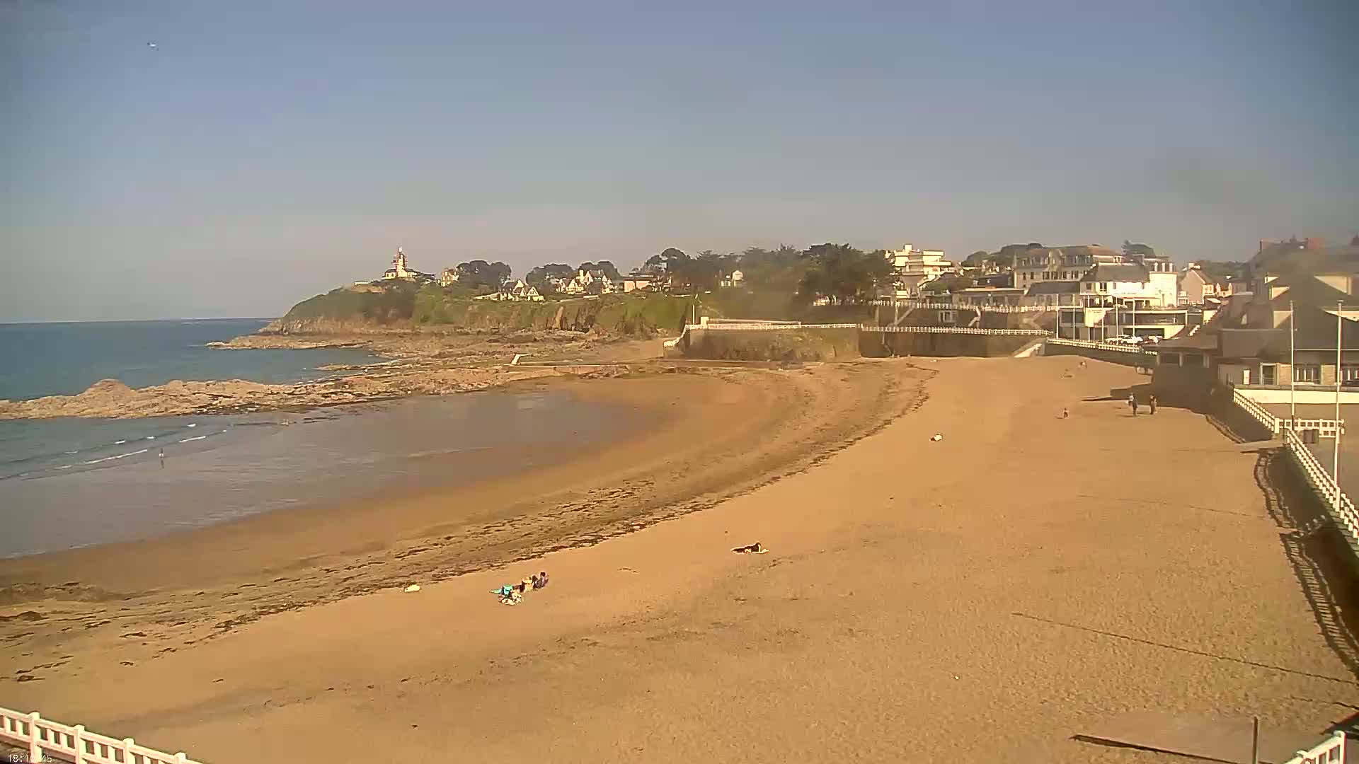A sandy beach curves around a calm bay, with buildings and a lighthouse visible on the headland in the distance under a clear sky.