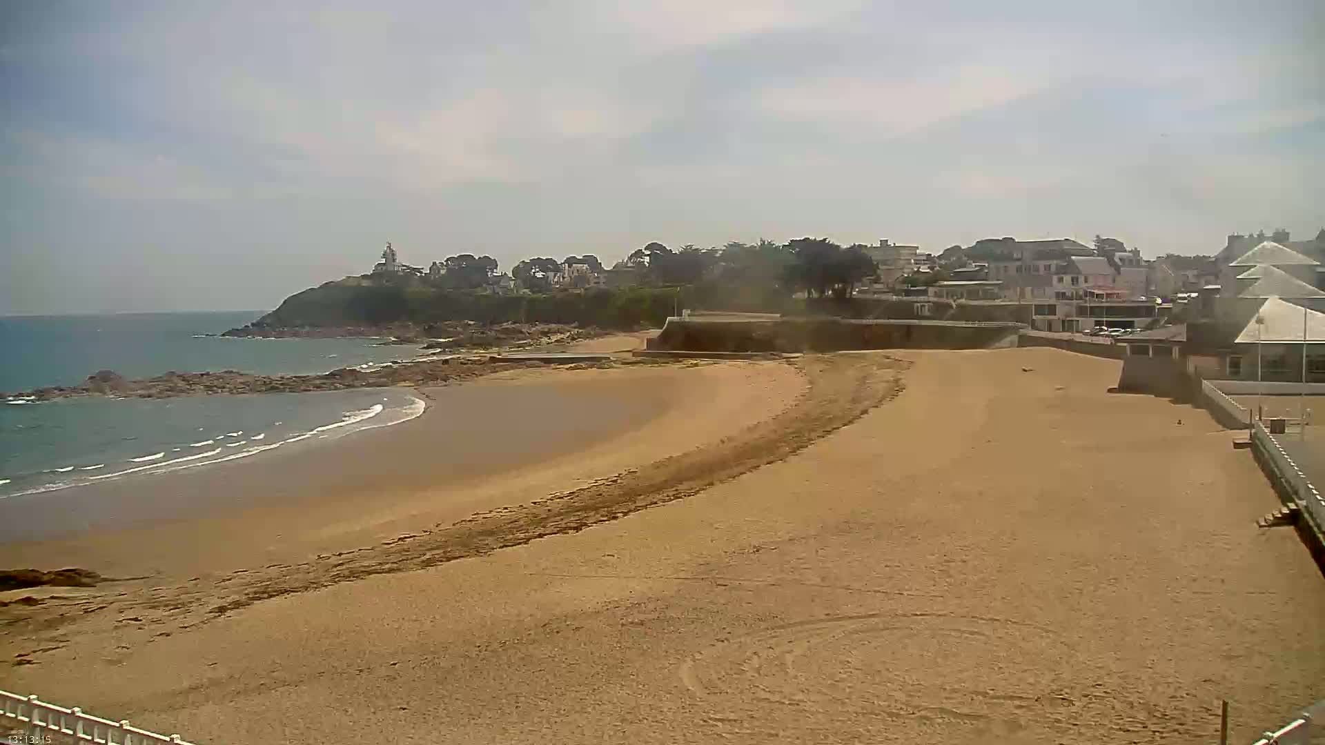 A sandy beach curves gently toward a rocky shoreline under a partly cloudy sky, with buildings and a lighthouse visible in the distance.