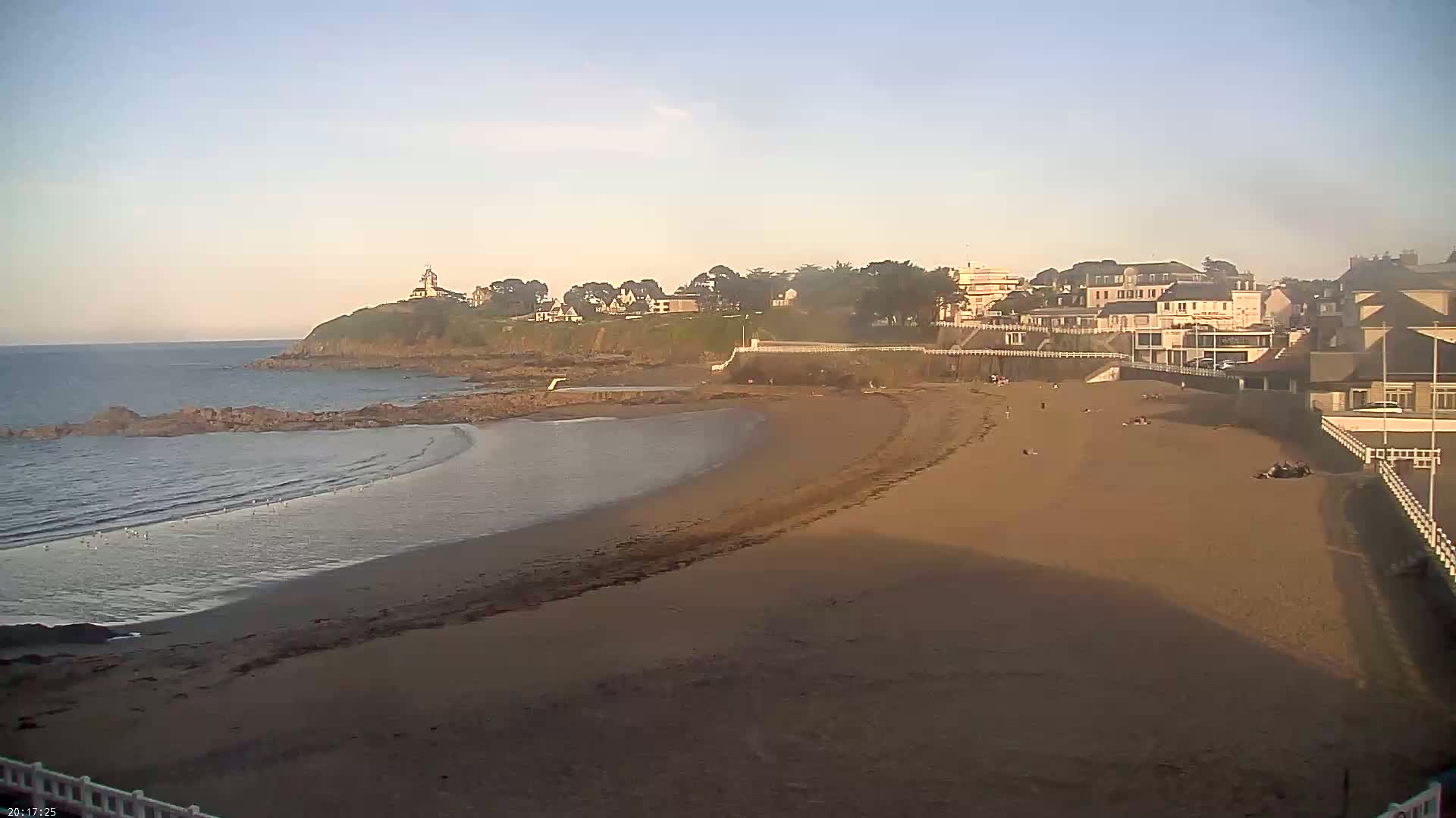 A sandy beach curves around a calm ocean, with a line of buildings and a small lighthouse visible on a cliff in the background under a clear, light-blue sky.