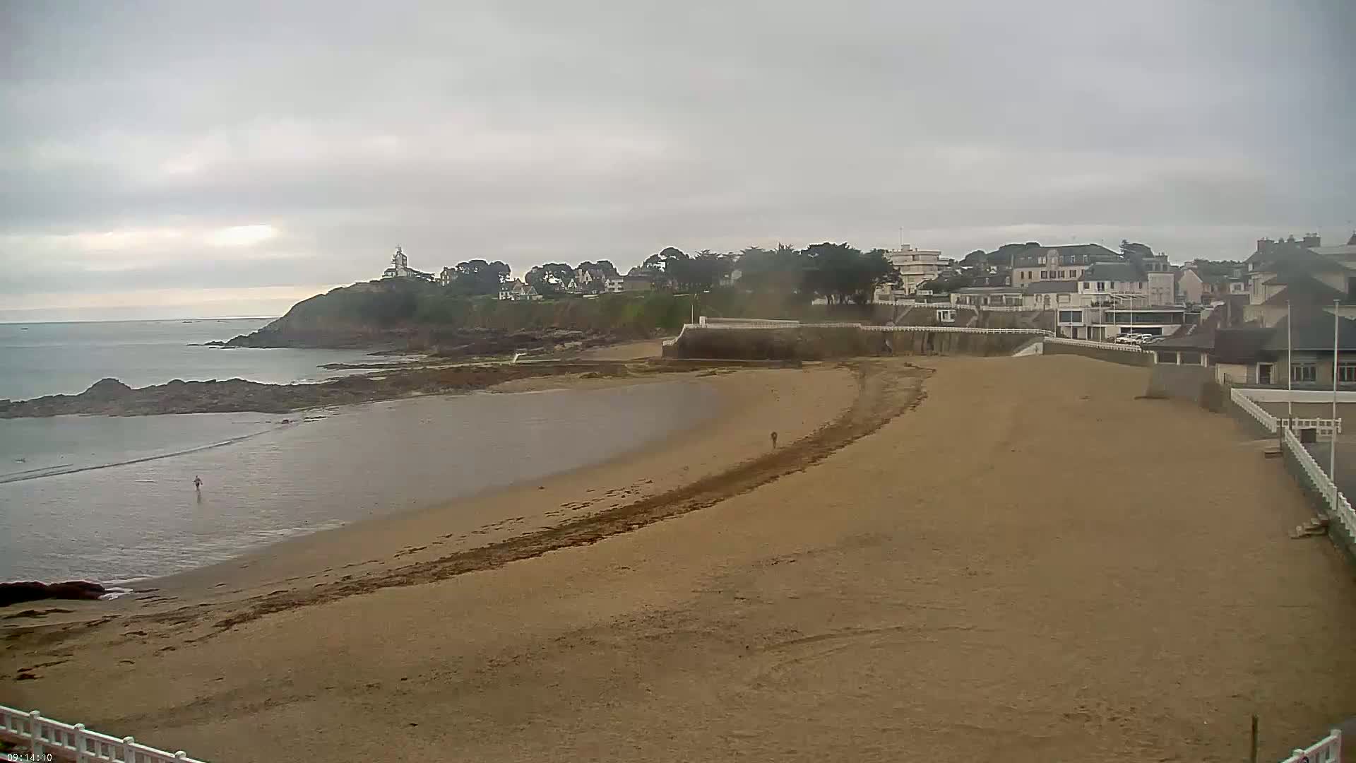 A sandy beach curves around a calm bay, under an overcast sky, towards a seaside town with houses and a lighthouse on a distant hill.